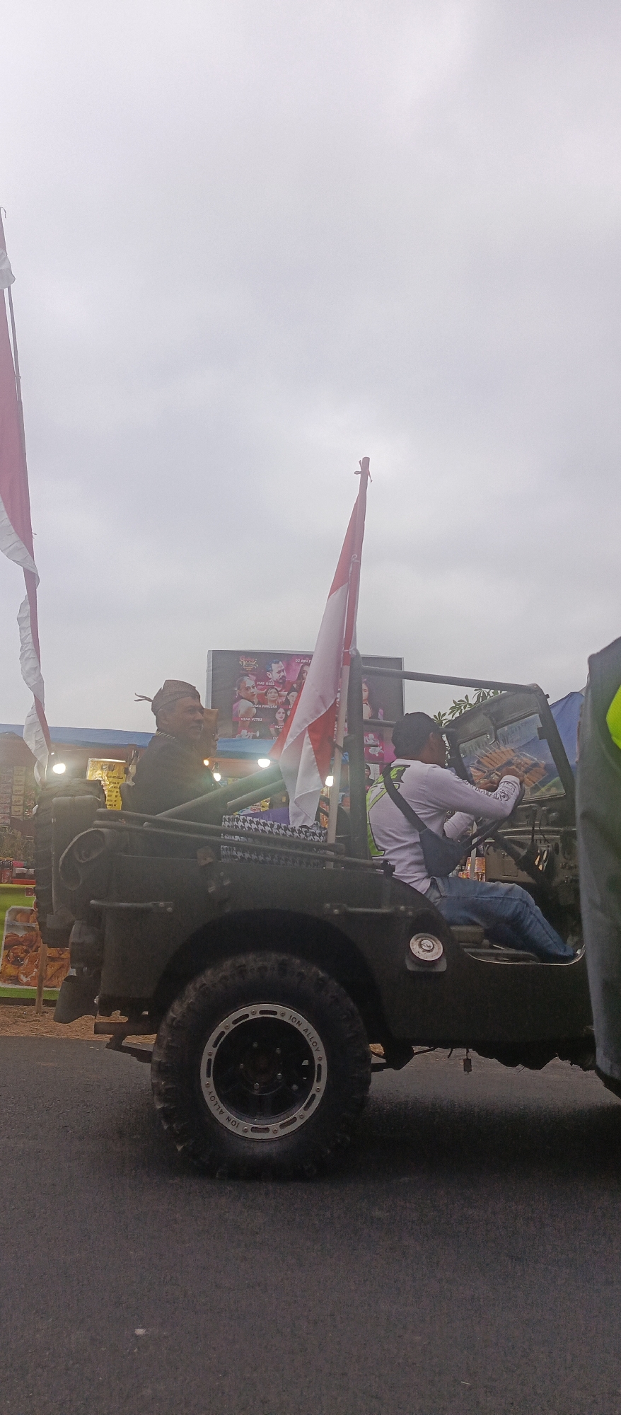 In Sumbersekar, Indonesia, an outdoor scene unfolds under an overcast sky. The foreground is dominated by the rear section of a dark green military-style jeep parked on an asphalt road. Its rugged tires, with "ION ALLOY" visible on the rim, hint at off-road capabilities. A small flag, split horizontally with red on top and white on the bottom, is affixed to a pole on the jeep's passenger side, flutters slightly.
Two men are seated in the jeep. The driver, wearing a white long-sleeved shirt and blue jeans, is actively steering. He appears to be holding a stack of what looks like bread or pastries in his right hand. On the driver's side, a second man, wearing a dark jacket and a traditional cap, looks towards the right of the frame, seemingly engaged in conversation or observation.
Behind the jeep, a large billboard displays colorful images of people, suggesting it might be an advertisement or event promotion. Two large flags, also red and white, are mounted on poles on either side of the jeep, further emphasizing national pride. The backdrop includes some structures and lighting, hinting at a public gathering or event. The overall atmosphere is one of a parade or festive procession.