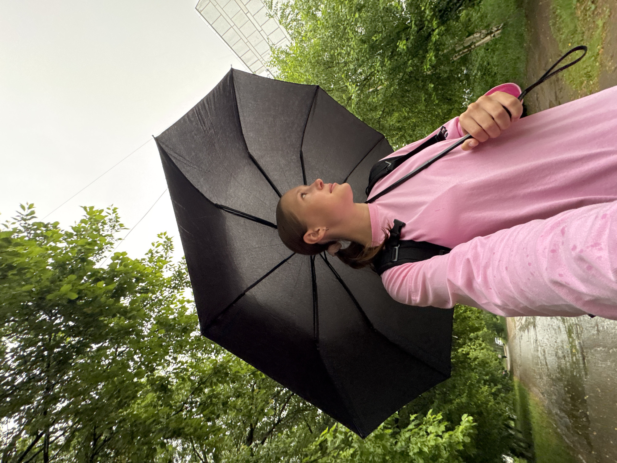 A young girl stands under a black umbrella outdoors. She is wearing a pink long-sleeved shirt and a black backpack. Her head is tilted upward as if she is looking at something in the sky. The background features lush green trees and a building with a grid-like facade visible through the foliage. A light-colored path or road runs horizontally through the scene on the right side. Water droplets can be seen on the sleeve of her shirt. The city of Chaikovsky, Russia, is not discernible in the scene.