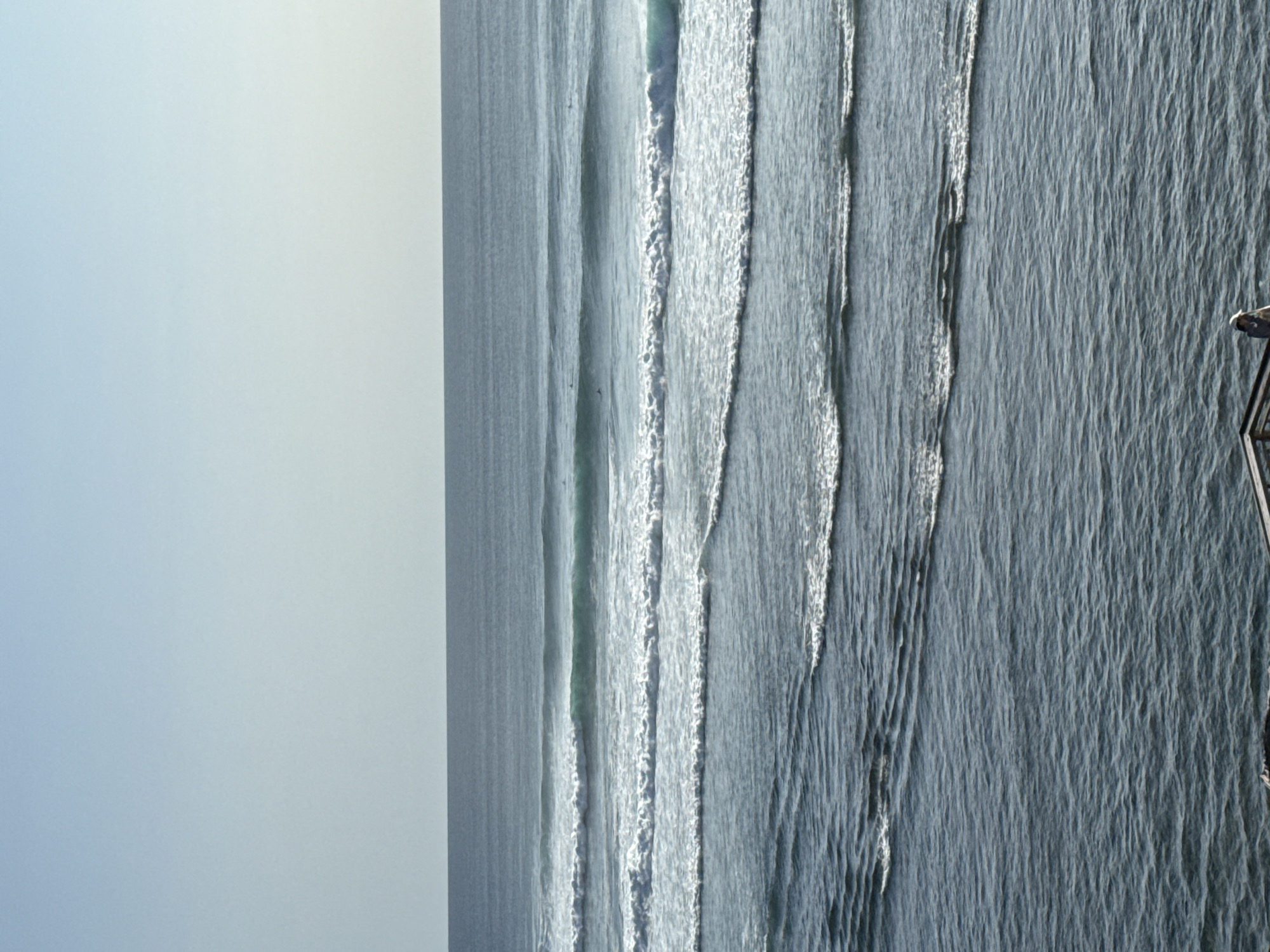 The image presents an aerial view of the ocean in Kuta Selatan, Indonesia. Consistent, uniform wave patterns dominate the water surface, with lines of white foam cresting the waves. A pier structure is barely visible at the right edge of the image. The sky is a uniform light gray or blue, suggesting an overcast or hazy day. No individuals are clearly identifiable within the water or on the pier. The general atmosphere is calm, with a focus on the repetitive motion of the ocean waves.