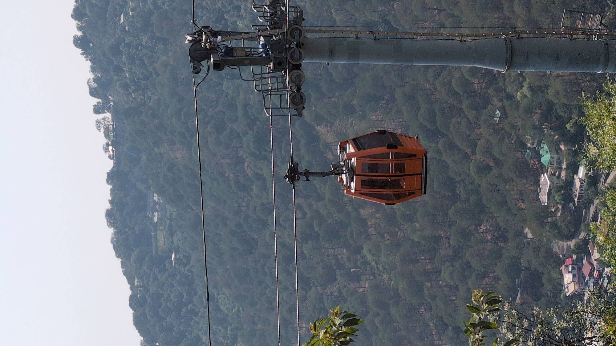 The image shows a cable car suspended on a ropeway, likely part of a transportation system in a mountainous region. The car is enclosed with a red-orange frame and appears to be traveling. Above the car is a support structure of the cableway, including a gray support tower and the wheel system used to carry the cable. The backdrop consists of a densely forested hillside dotted with buildings, likely the city of Dharamshala, India, due to the presence of pine trees on the mountain. The weather appears clear with a light blue sky in the background. The vegetation visible at the bottom of the image suggests a lush, natural environment.