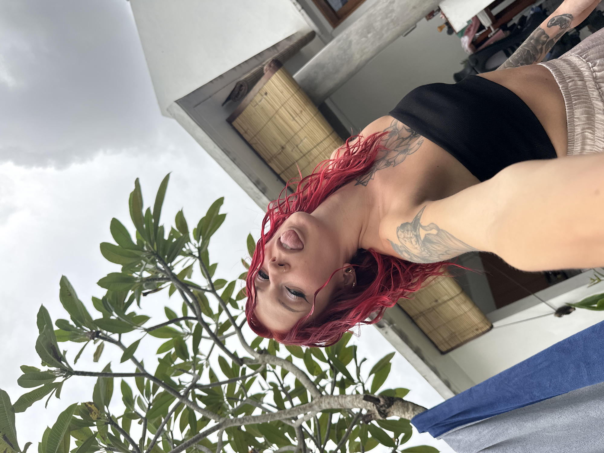 A woman with red hair and tattoos is the central subject of the image, sticking out her tongue in a playful expression. She is wearing a black top and light-colored shorts. The setting appears to be outdoors in North Kuta, Indonesia, suggested by the Balinese-style architecture partially visible in the background. A green tree occupies part of the lower left frame, and the sky is overcast. A blue and gray towel or cloth hangs in the foreground, possibly on a balcony or terrace.