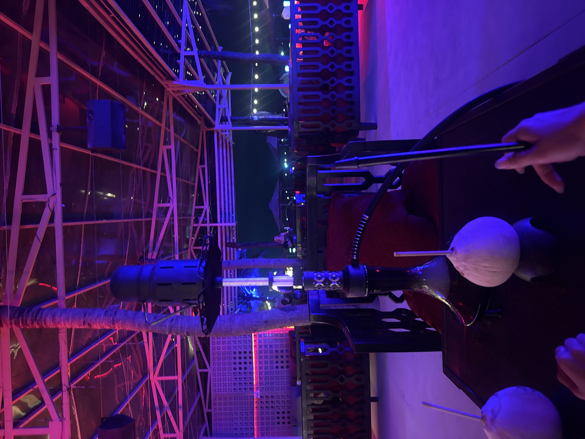The image shows a hookah with coconut bowls in a building in Đà Nẵng, Vietnam. The hookah is black with a glass base, and the coconut bowls are white and spherical. A person is holding the hookah pipe. The building has a metal frame and translucent walls, lit by purple and blue lights. There are decorative metal panels in the background.