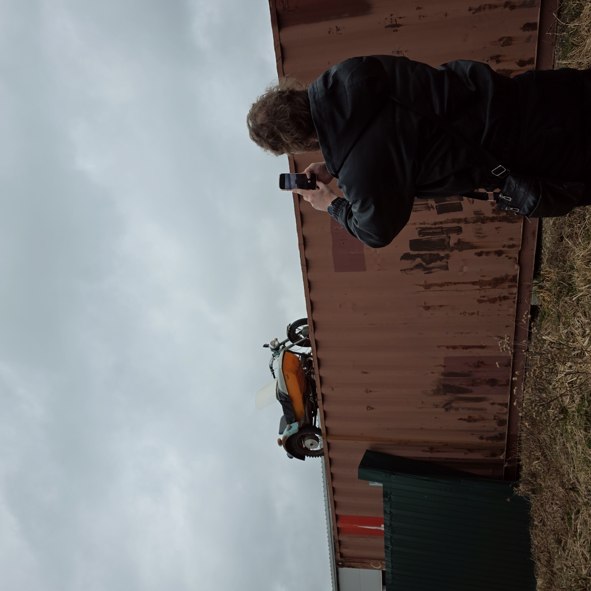 A person with curly brown hair is taking a photo with their smartphone. They are standing near a large, rusty, corrugated metal container. The person is wearing a black jacket and appears to have a black shoulder bag. In front of the container is an overturned motorcycle with an orange sidecar. The environment includes dry grass and an overcast sky. The exact location in the city of Volosovo, Russia, cannot be determined from this image.