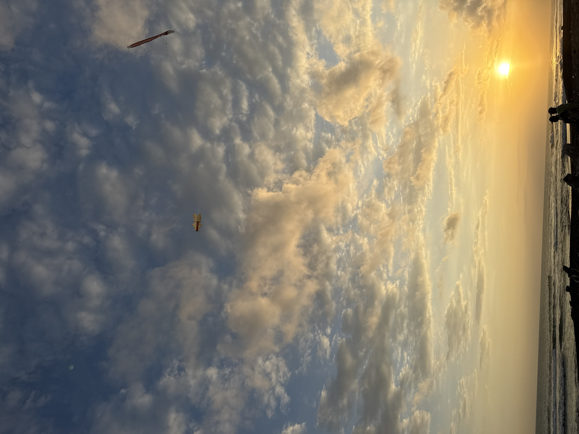 The image depicts a coastal scene in Unknown, Unknown, at sunset. The sky is filled with cumulus clouds, tinted with gold and orange hues from the setting sun, which is located near the horizon line at the top right of the image. The ocean is visible at the right edge, with small waves lapping at the shore. Two kites are visible in the sky: a longer, thinner kite in the top left, and a smaller, square kite in the lower left quadrant. Along the shoreline, possibly people, rocks, and debris are visible.