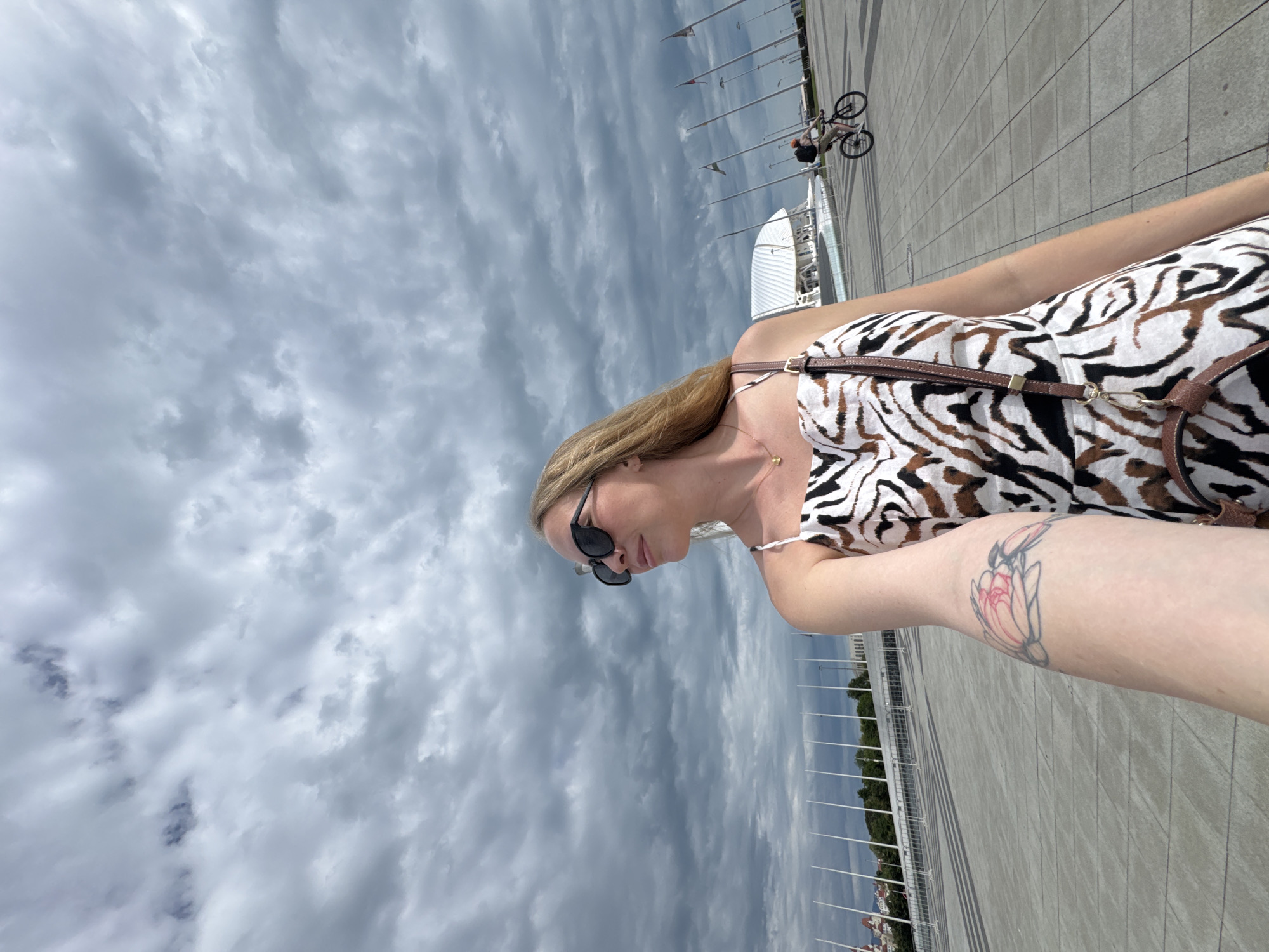 The image depicts a woman in a zebra-striped dress and sunglasses, standing on a paved surface with white poles running along the edge, likely a promenade. She has a tattoo on her arm. In the background, there is a cyclist and a white architectural structure. The sky is heavily clouded. The location appears to be the Olympic Park in Sirius, Russia, specifically near the Fisht Olympic Stadium.
