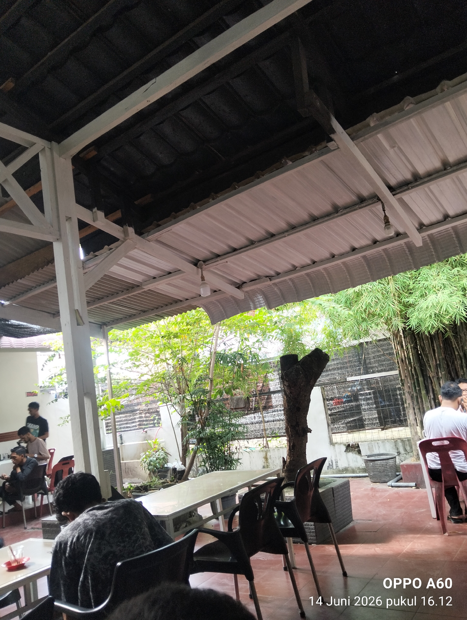 The image depicts an outdoor seating area, likely a cafe or restaurant, with several people present. The architecture suggests a tropical environment, with a corrugated metal roof supported by wooden beams. Green foliage, including bamboo, is visible in the background, indicating an outdoor or semi-outdoor setting.
In the foreground, a person wearing a grey patterned shirt is seated at a table, facing away from the camera. Another person in a black shirt is seated at a table to the left, also with their back to the camera. Further back, three individuals are standing near what appears to be an entrance or walkway. Two of them are wearing black t-shirts, and one is wearing a light brown t-shirt. One person is seated at a table on the far left. On the right side of the image, a person in a white t-shirt is seated at a red chair, facing towards the bamboo.
The seating area includes several tables and chairs. A long, narrow table is positioned centrally in the mid-ground. The floor is tiled in a reddish-brown color.
The timestamp overlay indicates "OPPO A60 14 Juni 2026 pukul 16.12", suggesting the image was taken on June 14, 2026, at 4:12 PM. The location is specified as Lhokseumawe, Indonesia, a city on the northern coast of Aceh province.