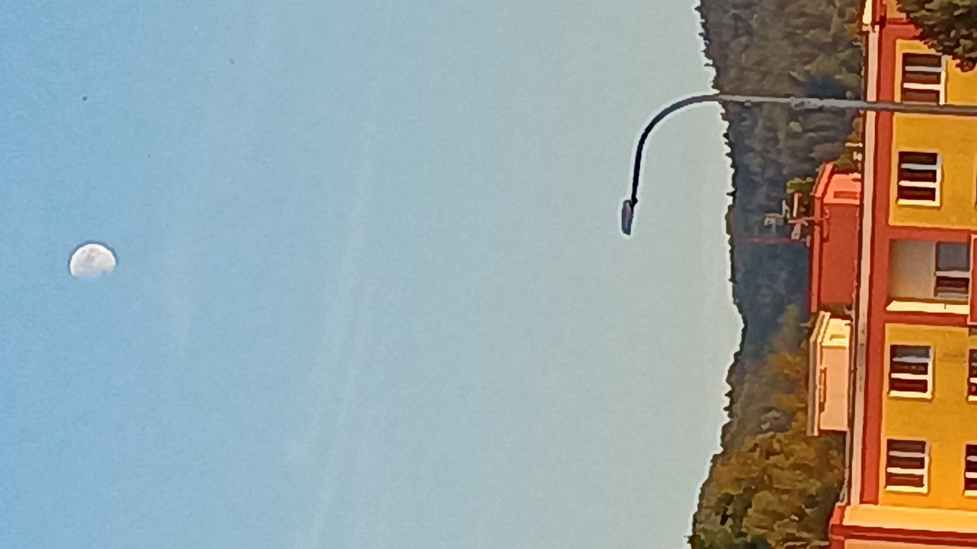 The image shows a view of the sky, a street lamp, and buildings in Sokolov, Czechia. The sky is clear and blue, with a gibbous moon visible in the upper portion of the frame. The moon is partially illuminated and appears to be in the daytime sky. A street lamp with a modern design curves into the frame from the right, its dark pole and fixture contrasting with the bright sky. Below the sky, a building with a yellow facade and red accents is visible. The building has multiple windows, some of which are dark and appear to be shuttered or tinted. A smaller, lighter-colored structure sits on top of the main building. Behind the buildings, a dense, green forest covers rolling hills. The overall impression is a daytime scene in a town or city, with natural elements of hills and forest in the background.