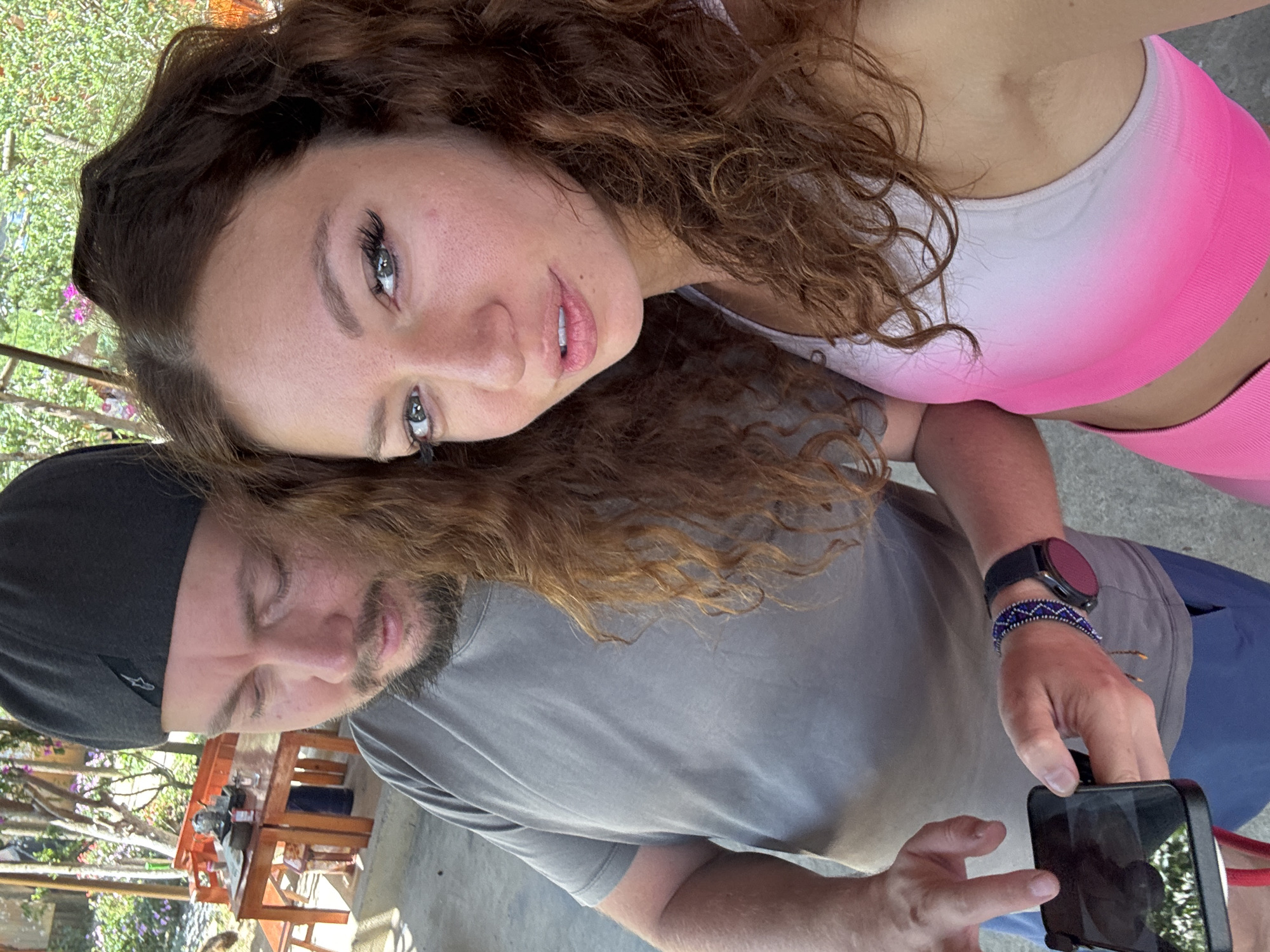 The image shows a woman with curly brown hair wearing a white and pink sports bra. She is positioned above a man with a beard, wearing a gray t-shirt and a black baseball cap. The man is holding a black smartphone, appearing to take a picture. The woman is looking at the camera with a slight smile. A smartwatch and a beaded bracelet are visible on the man's wrist. In the blurred background, there is outdoor furniture and greenery. The location is Bắc Hà, Vietnam.
