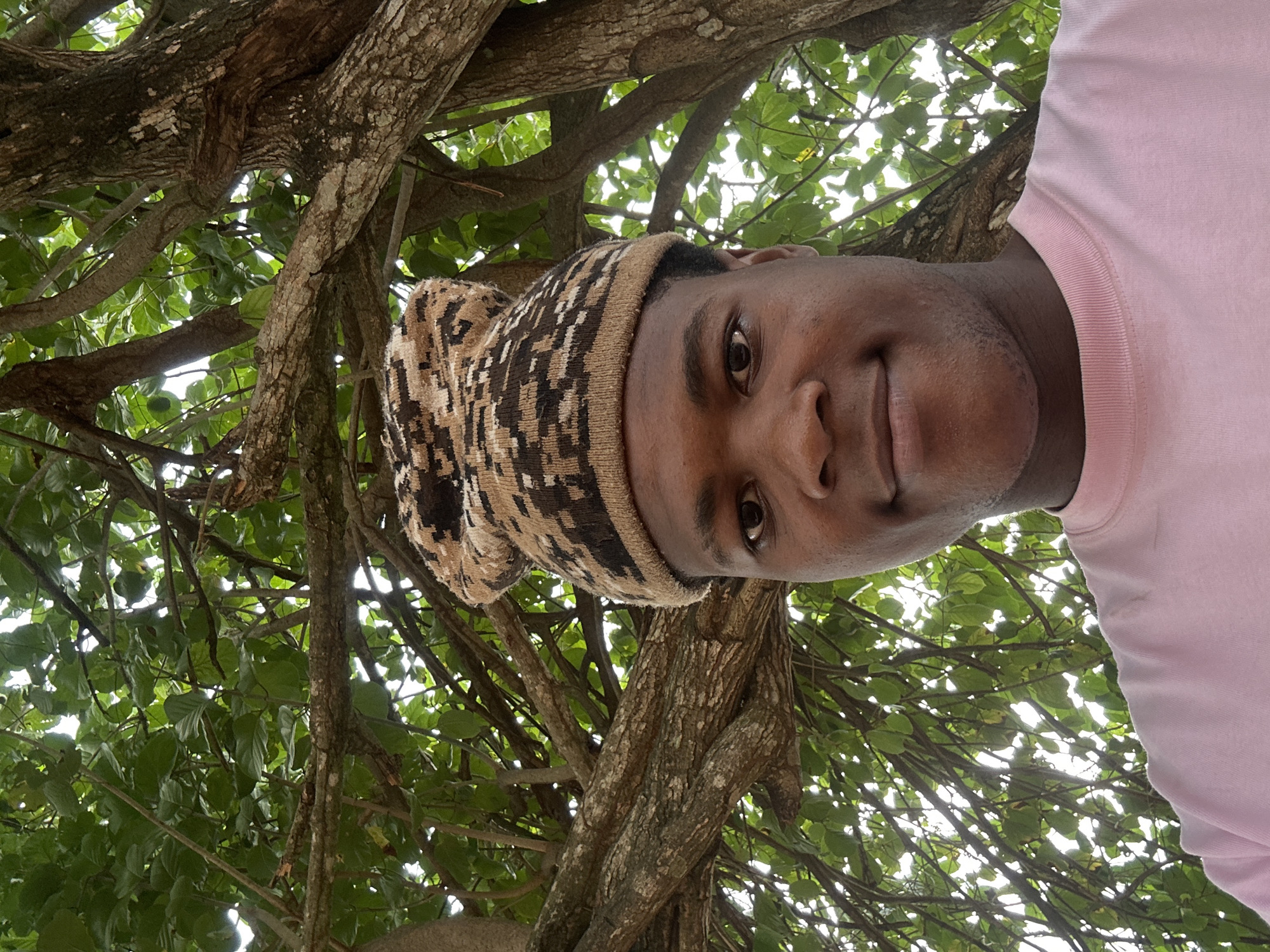A young Black male, seen from the chest up, is taking a selfie outdoors. He is wearing a pink crew-neck shirt and a patterned beanie hat in shades of brown, beige, and black. The hat has a folded brim and a pom-pom on top. He is looking directly at the camera with a slight smile. The background is dominated by the branches and green leaves of a large tree, with sunlight filtering through the foliage. The image appears to have been taken at a lower angle, looking up towards the subject and the tree. The location is identified as Ihiagwa, Nigeria.