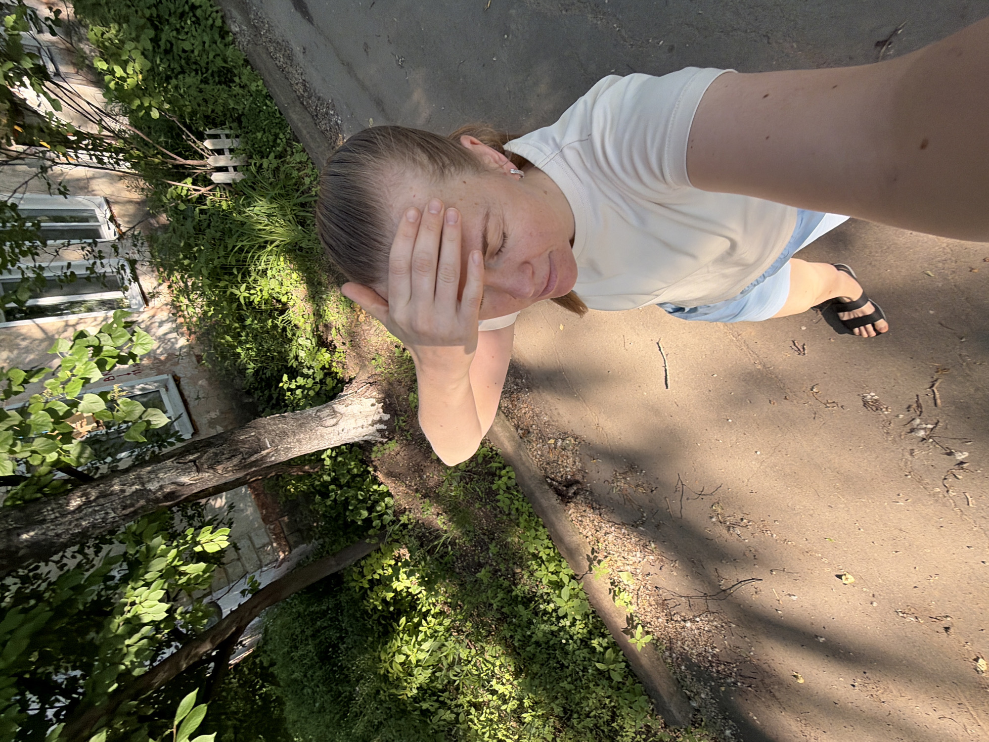 A woman is standing on a paved surface, possibly a sidewalk or road, with her right hand on her forehead, appearing to shield herself from the sun. She is wearing a light-colored t-shirt and light blue shorts. She is also wearing black sandals. The background includes a building with windows, trees, and dense green vegetation. There is a concrete curb separating the paved area from the vegetated area. The scene is illuminated by sunlight, creating shadows. The image appears to have been taken outdoors in a residential area. The presence of a building with visible windows and trees suggests an urban or suburban setting, consistent with a location in Khabarovsk, Russia.