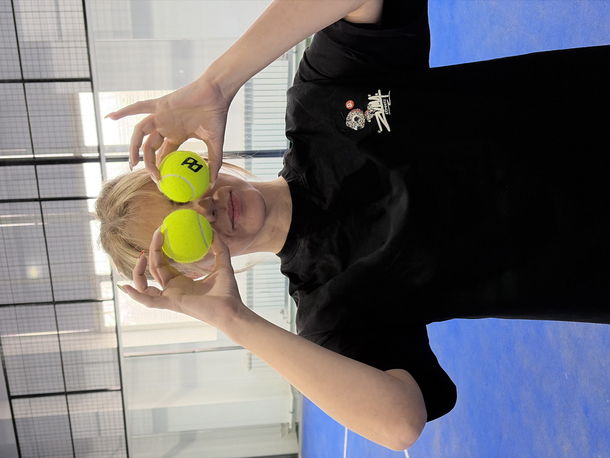 A young woman stands indoors, holding two yellow tennis balls up to her eyes as if they were spectacles. She wears a black t-shirt with a logo on the upper left side. Her blonde hair is visible behind the balls. The background includes a wall with a grid-like pattern and a floor with a blue surface. The location is in the city of Chaikovsky, Russia.