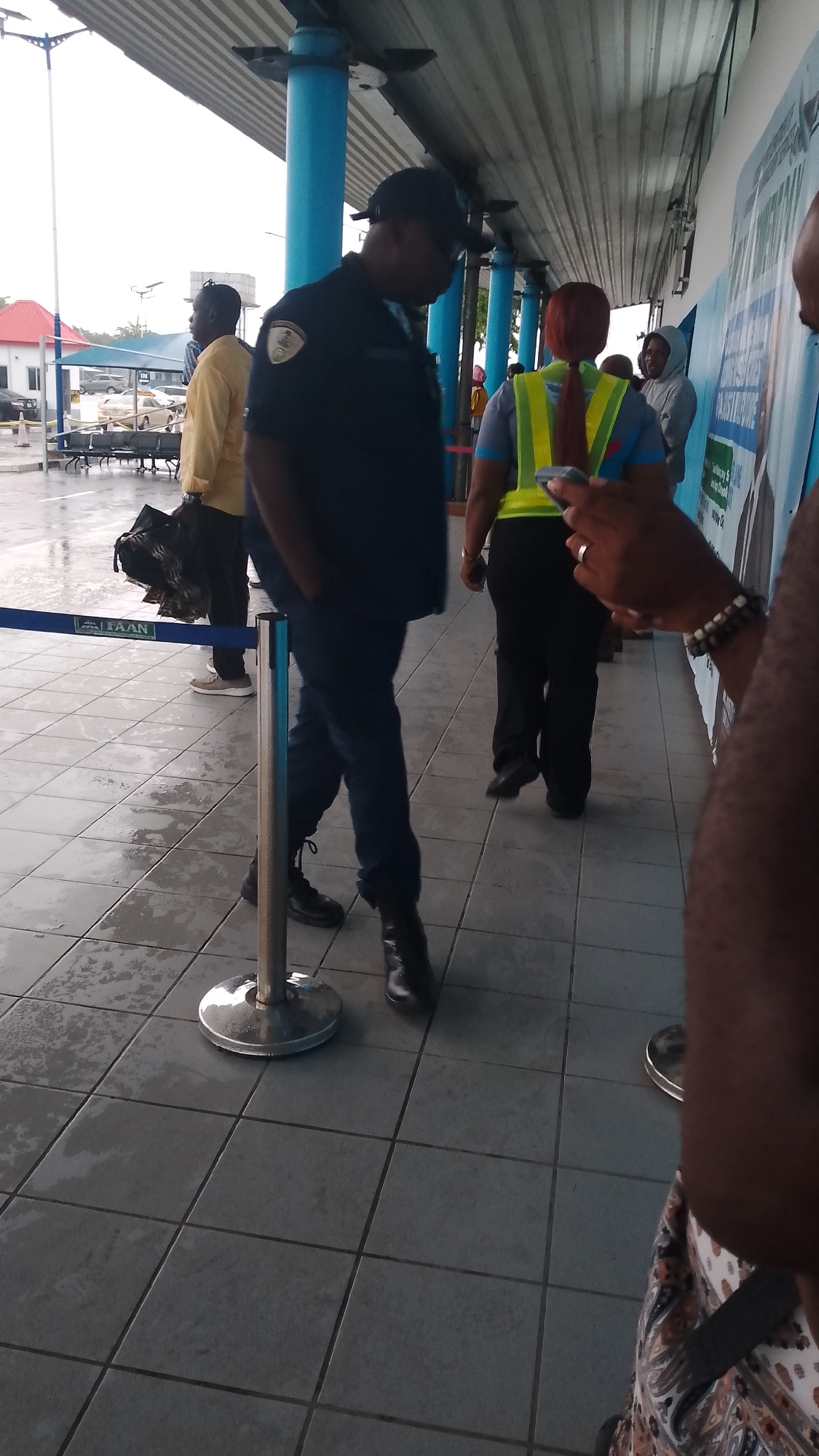 The image depicts an outdoor scene, likely at a transportation hub or public waiting area. The ground is tiled and appears wet, suggesting it may have rained recently. In the foreground, the legs of a person wearing dark pants and black boots are visible, standing next to a metal stanchion with a blue barrier tape that has "FAAN" printed on it.
Further back, a person in a dark uniform and a cap, likely a security guard, is walking away from the camera, with their hands in their pockets. To their right, a woman wearing a yellow and green reflective vest over a blue shirt and black pants is walking. Behind her, another person in a grey hooded sweatshirt is visible. To the left of the uniformed person, a man in a yellow shirt and dark pants is standing, holding a dark bag. In the background, there are buildings, some with red roofs, and what appears to be a water tower. The overall atmosphere is that of a functional, public space. The presence of the "FAAN" logo suggests this location may be related to airport or aviation services in Nigeria.