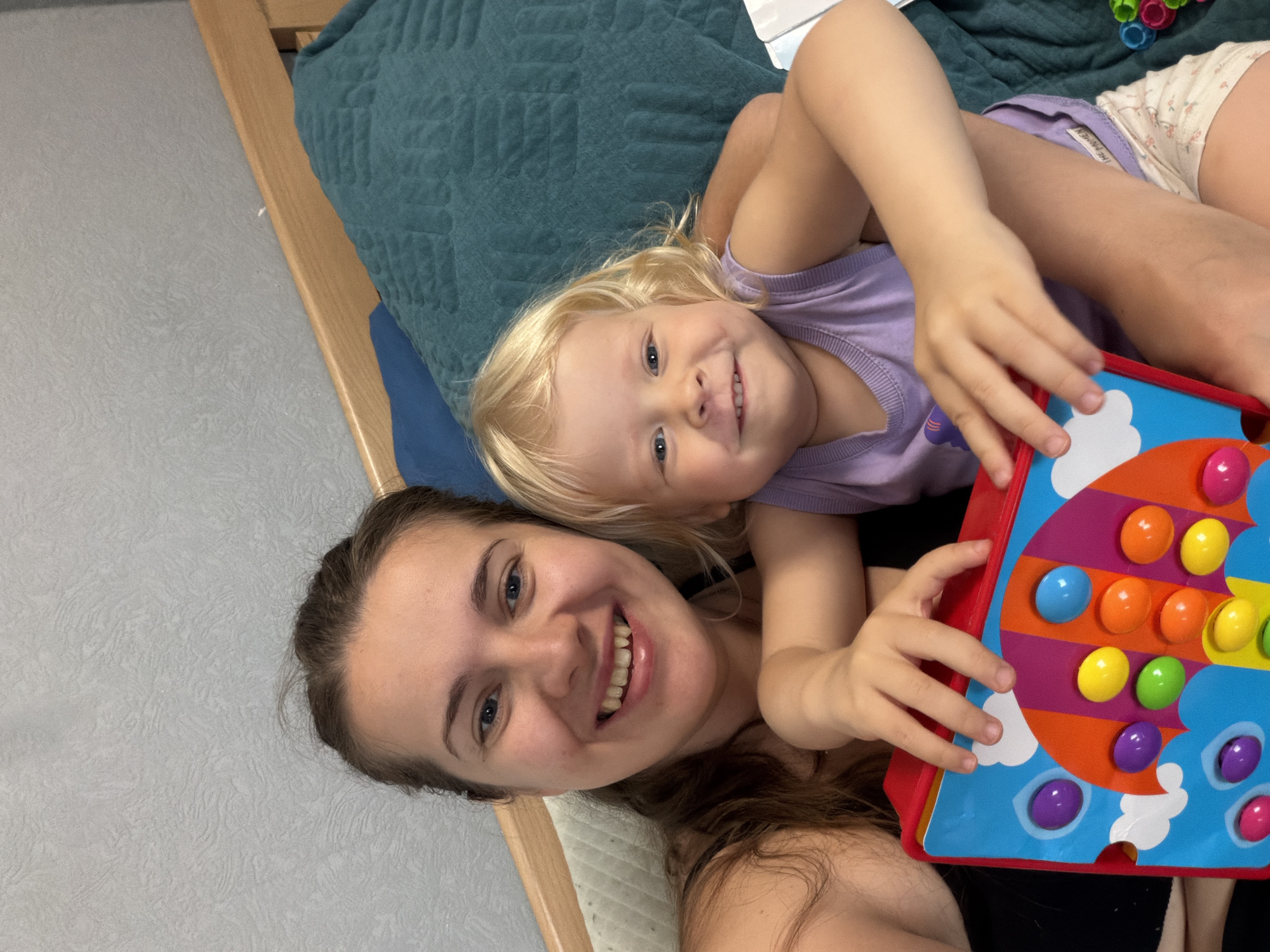 A selfie-style shot shows a woman and a young child lying down. The woman, with long brown hair and fair skin, is smiling broadly. The child, a blonde toddler wearing a purple tank top, is lying on the woman and also smiling. They are interacting with a colorful toy, which features buttons in various colors. The scene is indoors, with a bed and wall visible in the background. There is no information in the image to indicate this was taken in Sochi, Russia.