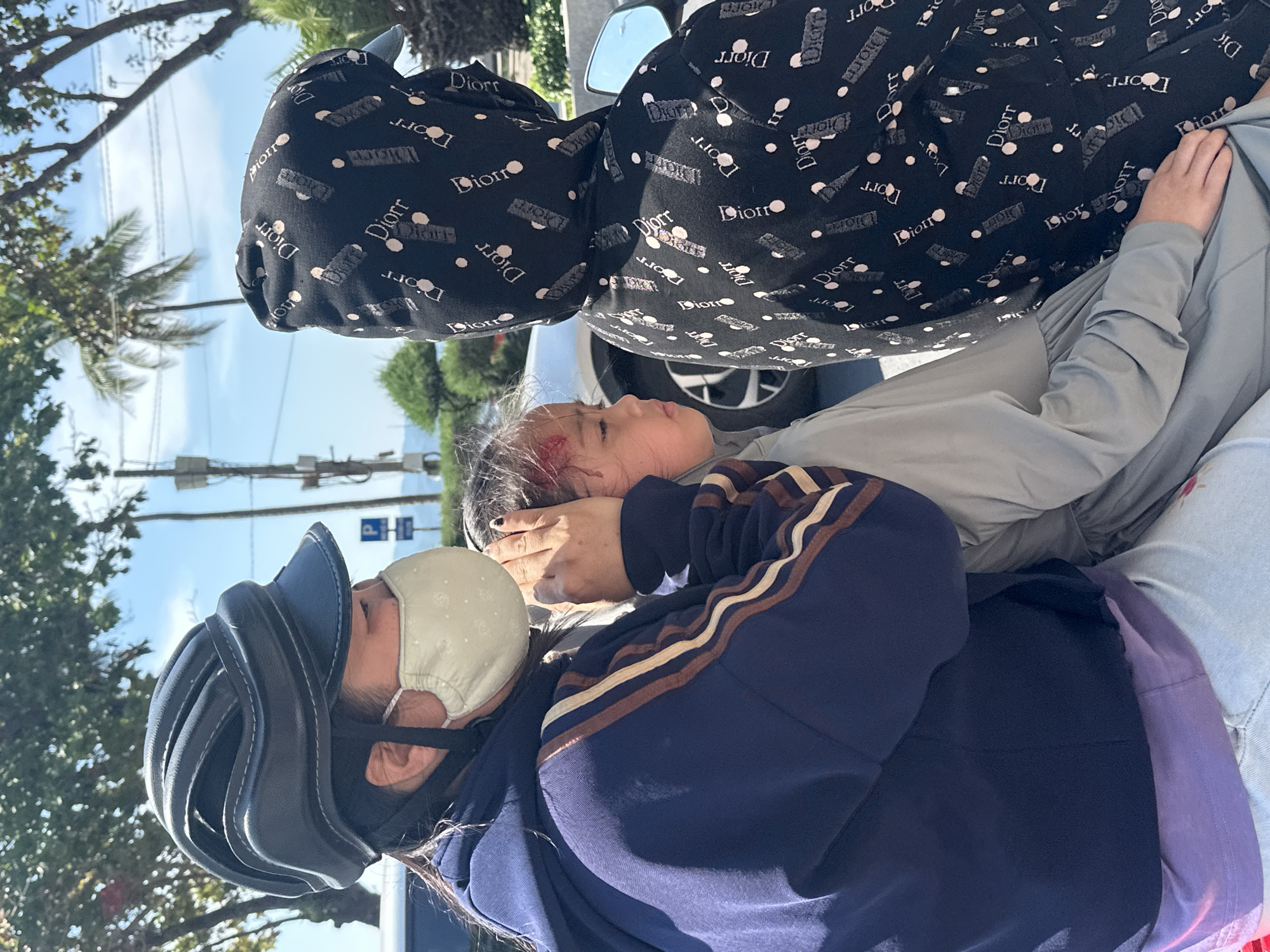 A concerned adult wearing a bicycle helmet, a light-colored face mask, and a dark navy blue sweatshirt with brown stripes on the shoulders, is supporting a young child who appears to have a head wound. The child is dressed in a light grey long-sleeved shirt and is lying on what appears to be a seat or lap, with another adult standing over them. The standing adult is wearing a black hooded sweatshirt with the word "Dior" printed repeatedly on it. The scene seems to be outdoors, potentially in a city street setting, possibly Nha Trang Ward, Vietnam, suggested by the image's origin. Trees, power lines, and possibly a vehicle are visible in the background.