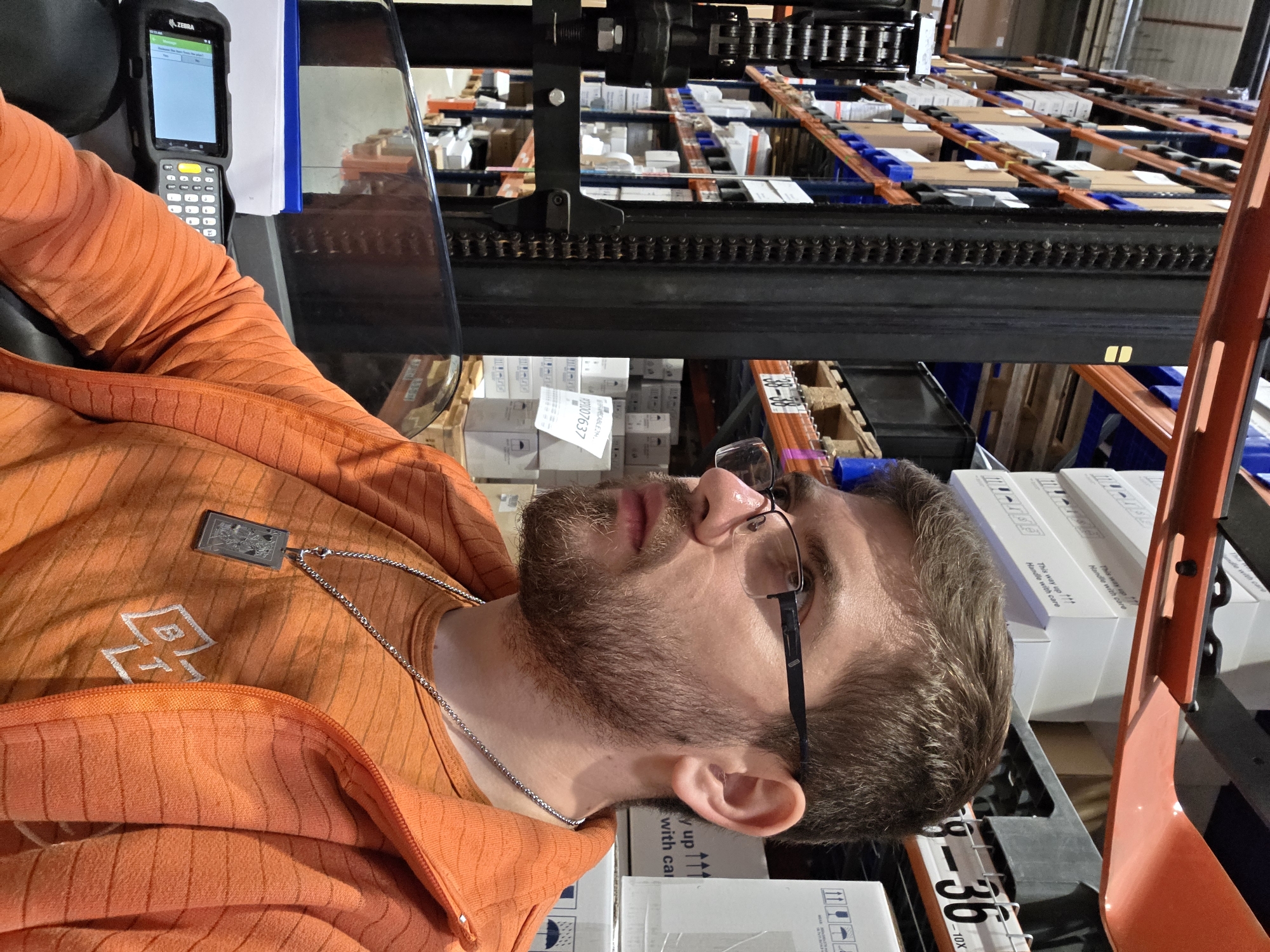 A man with a beard and glasses is lying on boxes in what appears to be a warehouse or storage facility. He is wearing an orange jacket and a silver chain with a pendant. A Zebra mobile device is next to him. Pallet racking filled with boxes is visible in the background. The boxes on which he's lying have shipping labels with text including "This way up," "Handle with care," and serial number/model text such as "-36" and "-10X." There's also shipping information (such as "MECABLEZM" and "POP1007637") visible on box labels. There is nothing indicating this scene is in Plovdiv, Bulgaria.
