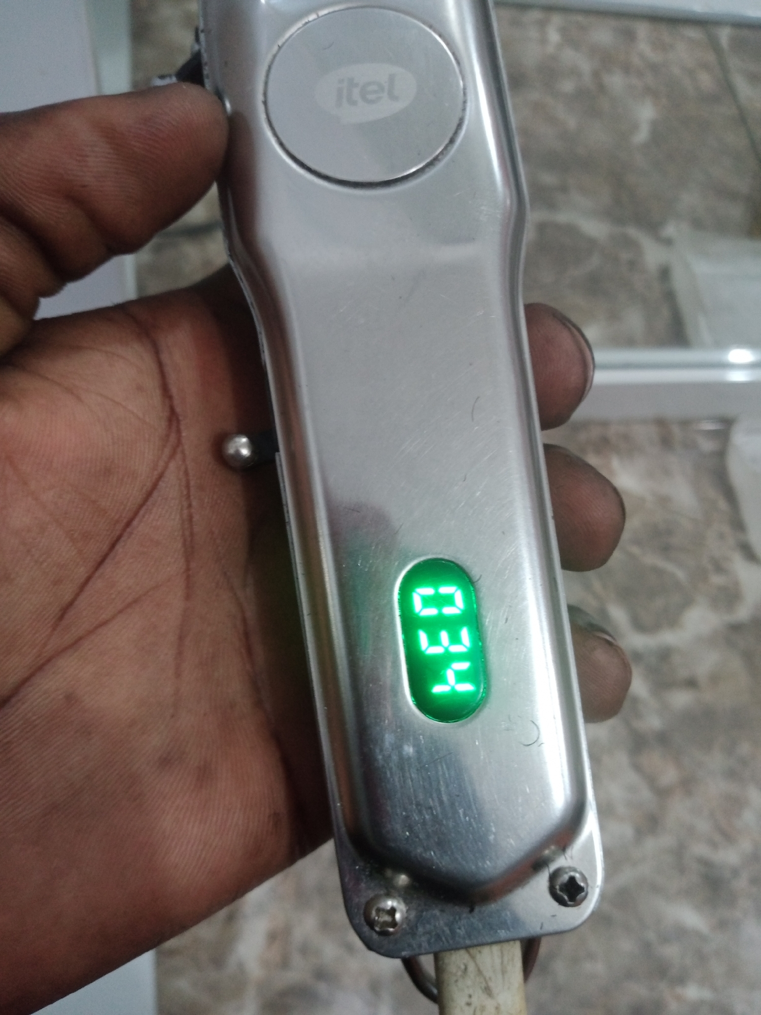 In Jalingo, Nigeria, a close-up, eye-level, portrait-oriented shot focuses on a silver electric hair clipper held by a person's hand. The hand, with visible lines and texture, is positioned on the left side of the frame, obscuring part of the clipper. The clipper, a prominent subject, is silver with a circular button near the top displaying the brand "itel" in grey text. Below this, a green LED digital display shows the numbers "0.34". The background is blurred, showing a mottled grey surface and hints of a light-colored vertical structure, possibly a wall or shelf, and a white frame structure in the upper right. The lighting appears to be indoor ambient light, casting soft shadows.