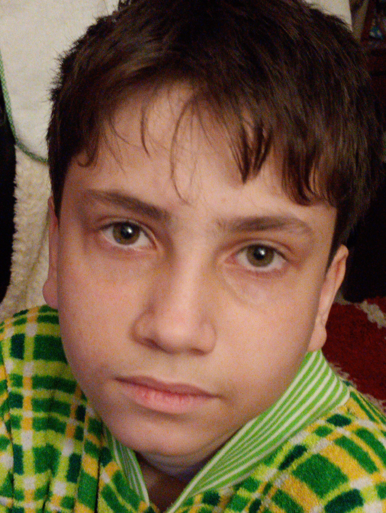 The image shows a close-up portrait of a young boy with brown hair, looking directly at the camera. He is wearing a green and yellow checkered shirt with a striped collar. The background is blurred, with some hints of a light-colored fabric on the left and a dark, possibly red, patterned fabric on the right. There is no discernible information about the location in Horishni Plavni, Ukraine, within the image itself.