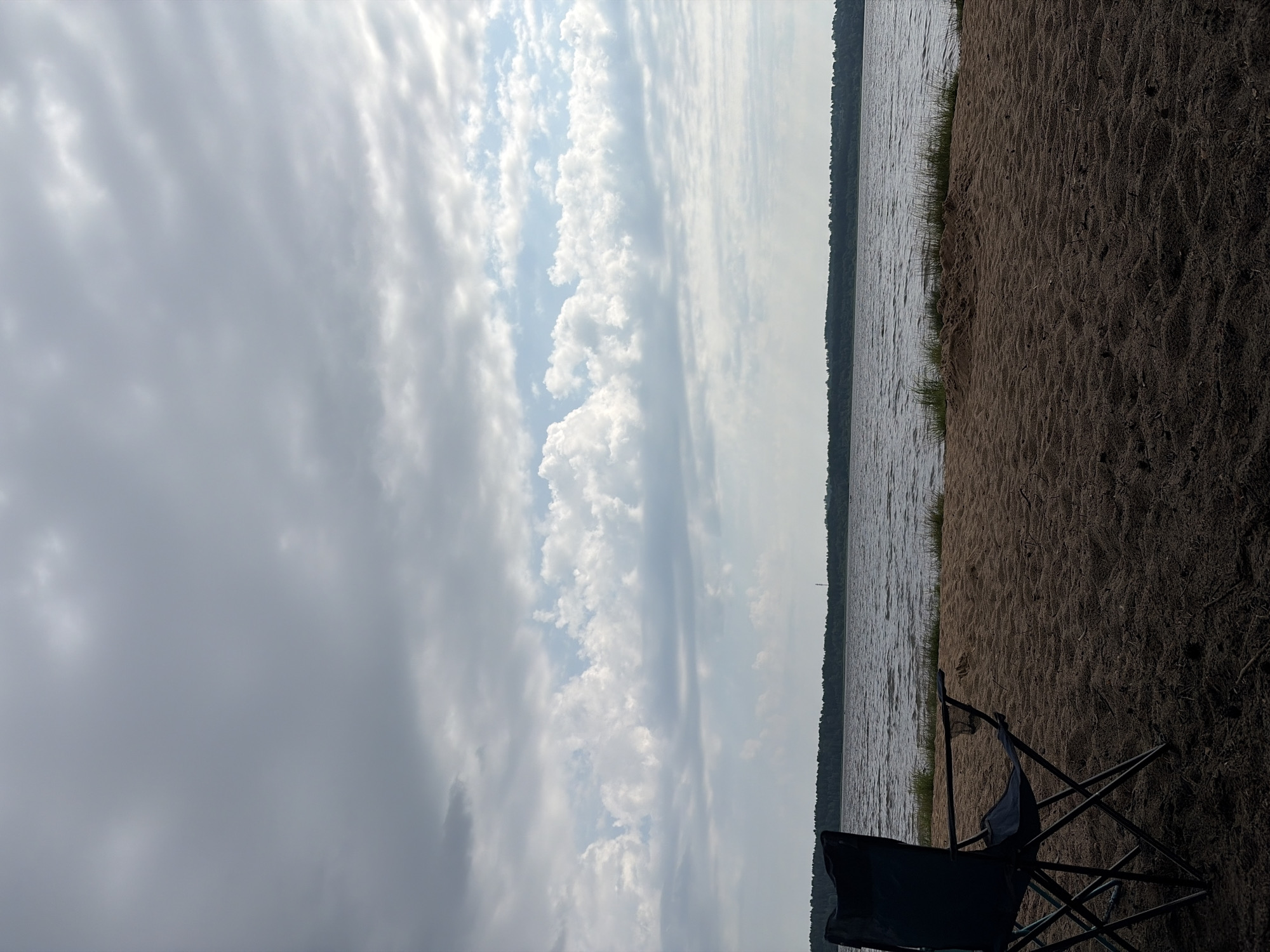 This is a photograph of a sandy beach on the shore of a large body of water, likely a lake or river, with a dark, forest-covered horizon line in the distance. The sky is filled with a mix of gray and white clouds, with some patches of blue visible. In the foreground, a dark blue camping chair is positioned on the sand, facing the water. The sand is coarse and appears somewhat damp, with small pieces of debris scattered across it. The water is rippled, suggesting a light breeze. The overall scene depicts a serene, natural environment, possibly a recreational area in Vaskelovo, Russia. There are no people visible in the image.