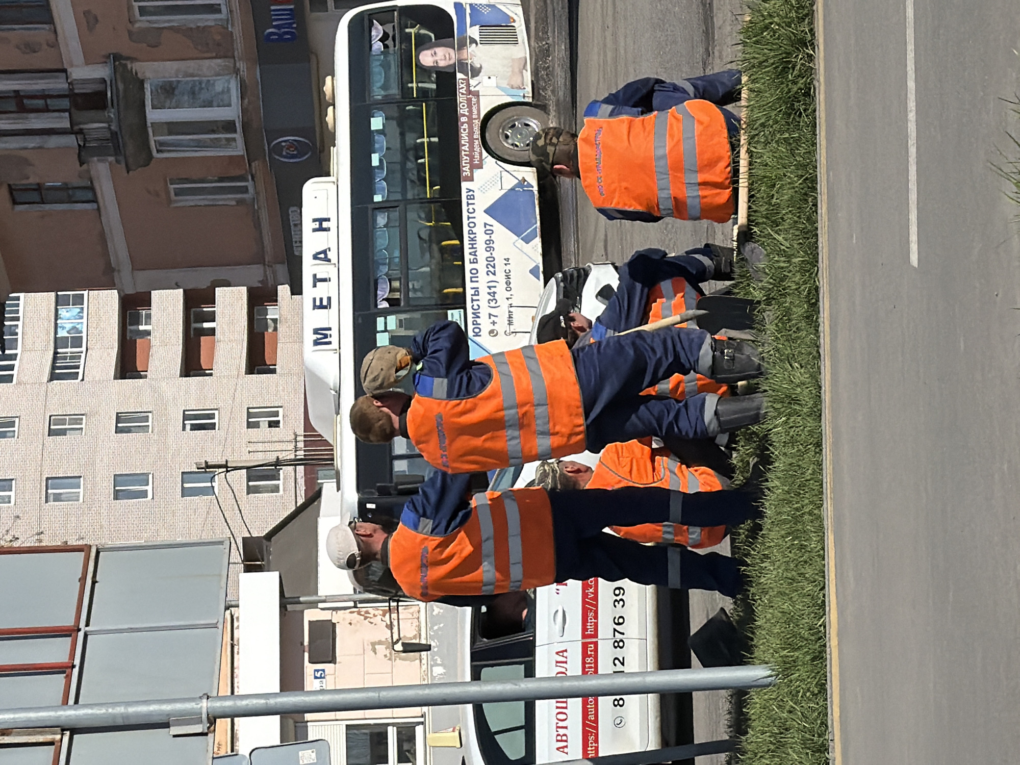 The image shows four road workers in Votkinsk, Russia, wearing orange vests and blue uniforms, working on the edge of a road with grass nearby. A bus with advertising for bankruptcy lawyers and the word "METAH" is present in the background. A car from a driving school is also parked nearby. Buildings are visible in the background.