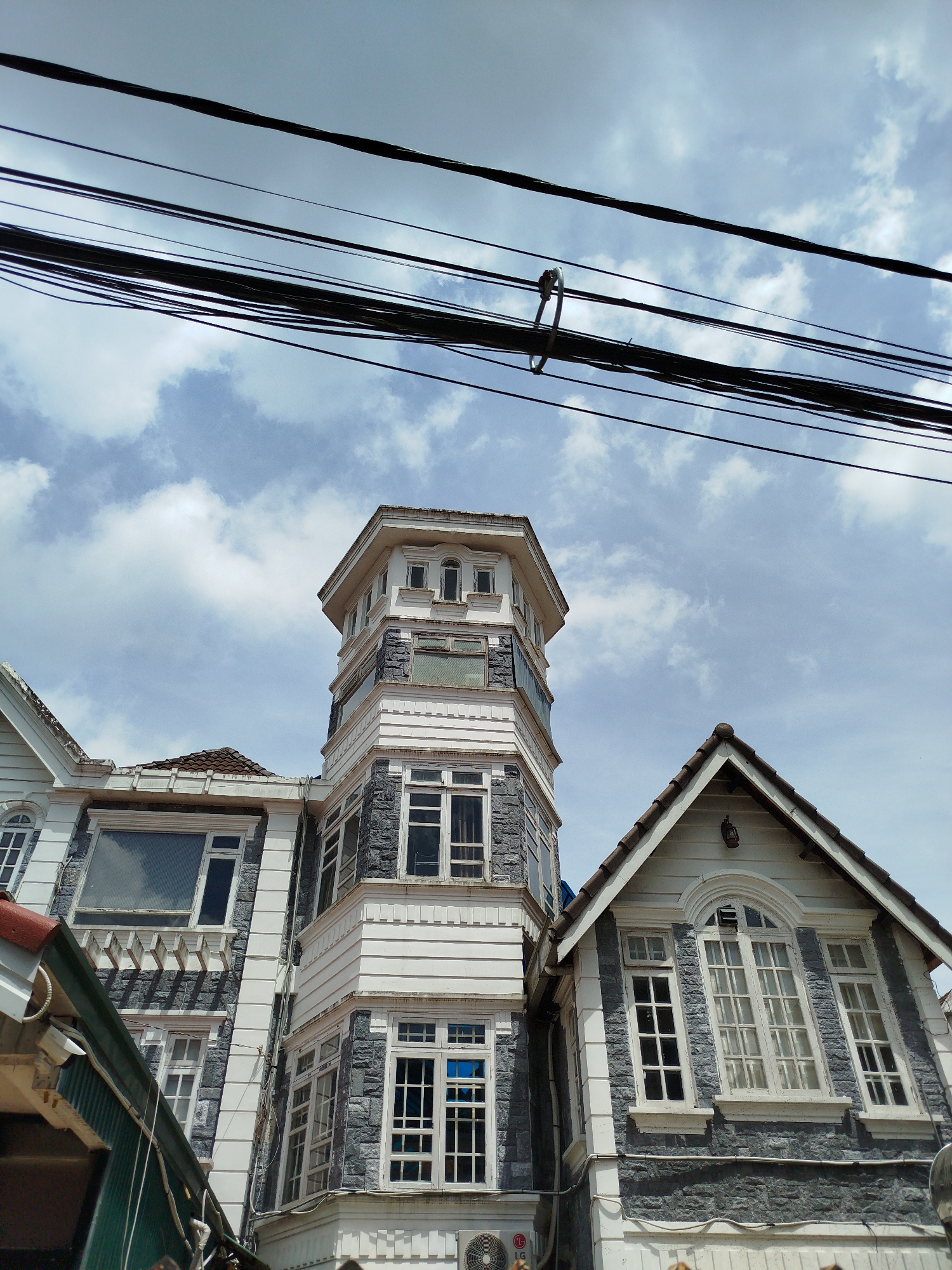 The image shows a multi-story building with a unique architectural style, possibly in Da Lat, Vietnam, given the context. The building features a prominent octagonal tower with white trim and stone-like siding, topped with a lighter-colored section that has multiple windows. To the right of the tower, a gabled section of the building is visible, also with white trim and stone-like siding, featuring a large arched window with multiple panes. Several other windows are visible on different levels of the building, most of them framed in white. Overhanging the scene are several black electrical wires, some appearing to be suspended from a metal ring. The sky above is partly cloudy with a bright, daytime light. The overall impression is a residential or commercial building with a blend of European and local architectural influences. No people are visible in the frame.