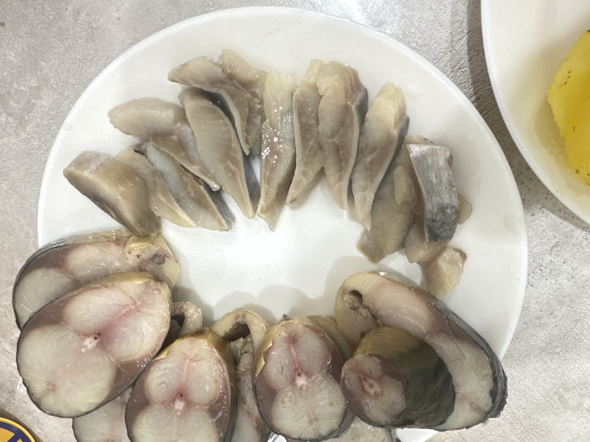 A white plate is centrally located in the image and sits on a light-colored, textured surface. On the plate, slices of fish are arranged in two groups. The top group consists of long, diagonally cut fillets with a silvery-gray skin and light-colored flesh. The lower group is composed of circular fish slices, showcasing a marbled pattern of light and dark meat, with darker skin. Another white plate with a yellow object is slightly visible in the upper-right corner. The image was taken in Chaikovsky, Russia.