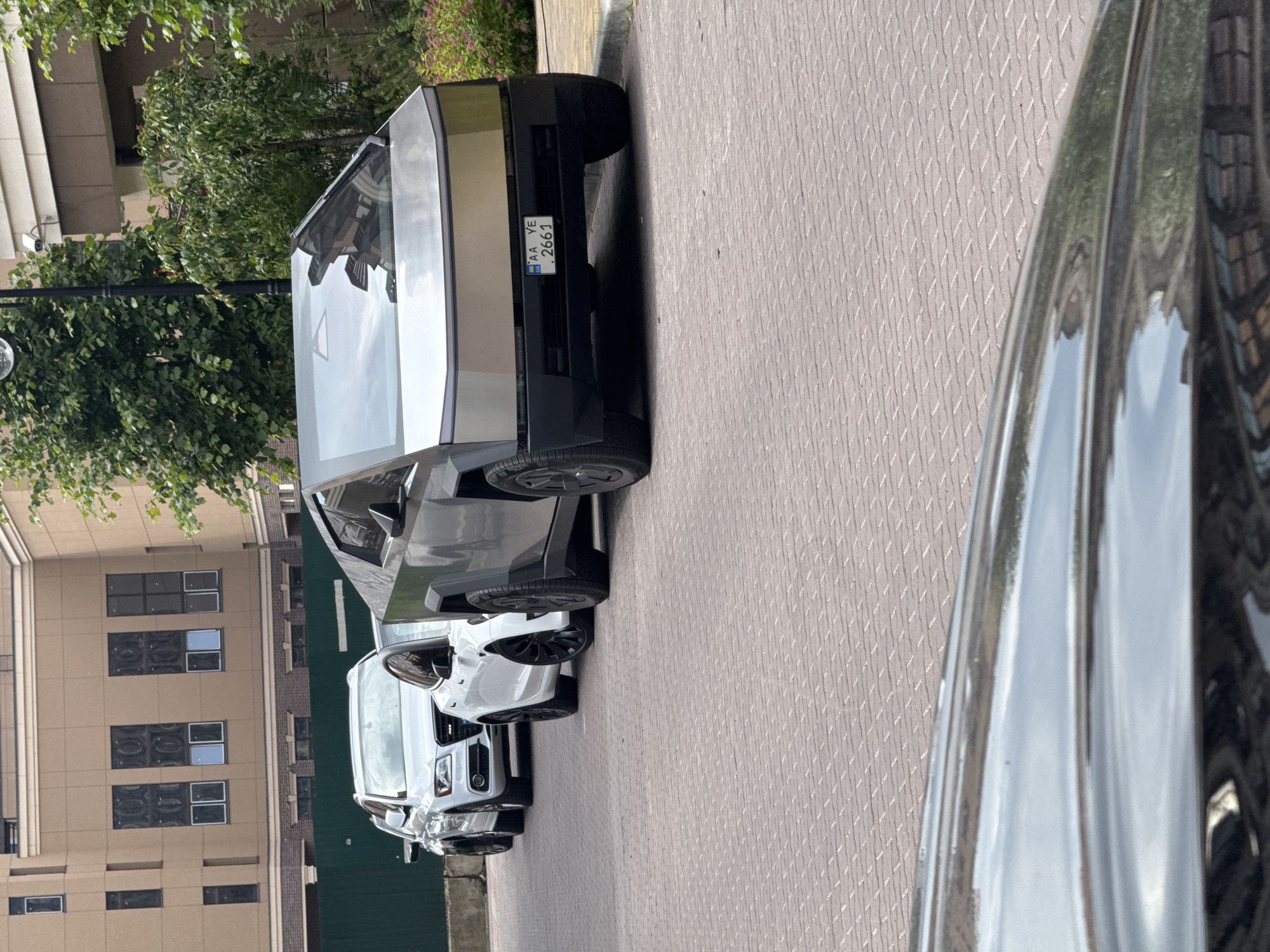 The image depicts an outdoor urban environment in Sofiivska Borshchahivka, Ukraine, during daylight hours. Three vehicles are parked in a line on a light-colored paver surface, which exhibits a diagonal herringbone pattern.
In the immediate foreground, positioned to the right, is a silver-grey Tesla Cybertruck. Its angular, unpainted stainless steel body reflects the surrounding environment. The front fascia is visible, featuring a Ukrainian license plate displaying "UA AA YE 2661". Black wheel arches and multi-spoke wheels with visible tire tread are present. Reflections of trees and buildings are apparent on the windshield and body panels.
Behind the Cybertruck, slightly to its left, is a white Tesla sedan, likely a Model 3. Its black multi-spoke wheels with red brake calipers are discernible.
Further back, behind the white Tesla and to the left, is a white Audi SUV, identifiable by its prominent front grille and headlights, resembling an Audi Q5 model.
The vehicles are parked alongside a grey curb, beyond which a narrow strip of ground with some sparse vegetation is visible. In the background, a multi-story building constructed of light brown (tan) material with a darker brick base is present. The building features multiple rectangular windows, some of which exhibit internal patterns or reflections. A dark green privacy fence runs horizontally across the mid-ground behind the Audi. Lush green deciduous trees and bushes are scattered throughout the background, providing foliage. A black lamppost with a spherical light fixture and a white CCTV camera is partially visible in the upper right background, amidst the trees. No individuals are visible in the scene.