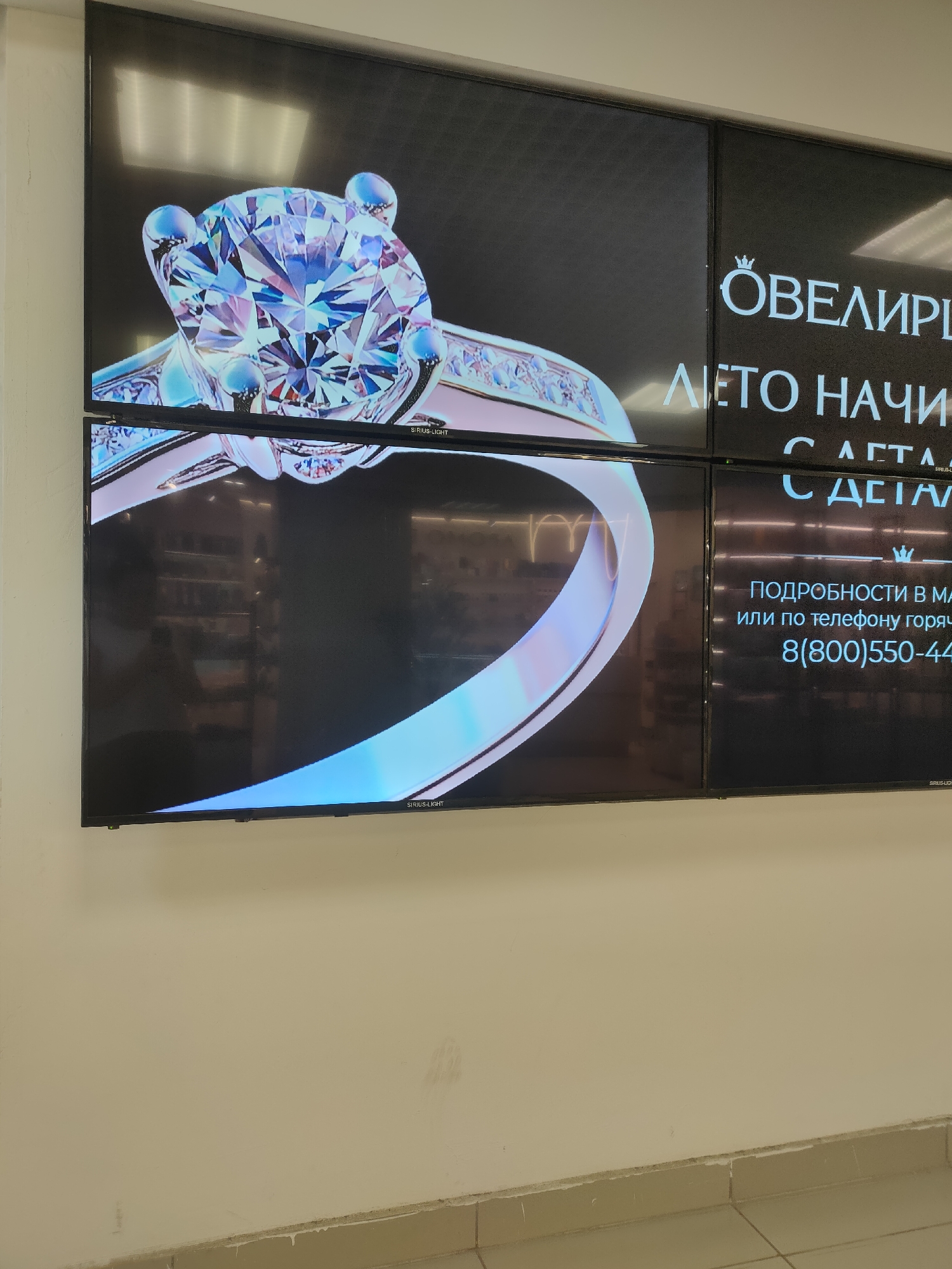 A video display screen shows a diamond ring on a black background. Text in Russian appears on the screen, including "ЛЕТО НАЧИНАЕТСЯ С ДЕТАЛЯМИ" which translates to "Summer begins with details" and "ПОДРОБНОСТИ В МАГАЗИНЕ или по телефону горячей линии 8(800)550-4400", which translates to "Details in the store or by calling the hotline 8(800)550-4400". The screen is mounted on a light-colored wall in what appears to be a commercial setting. The image was taken in Oktyabrsky, Russia.