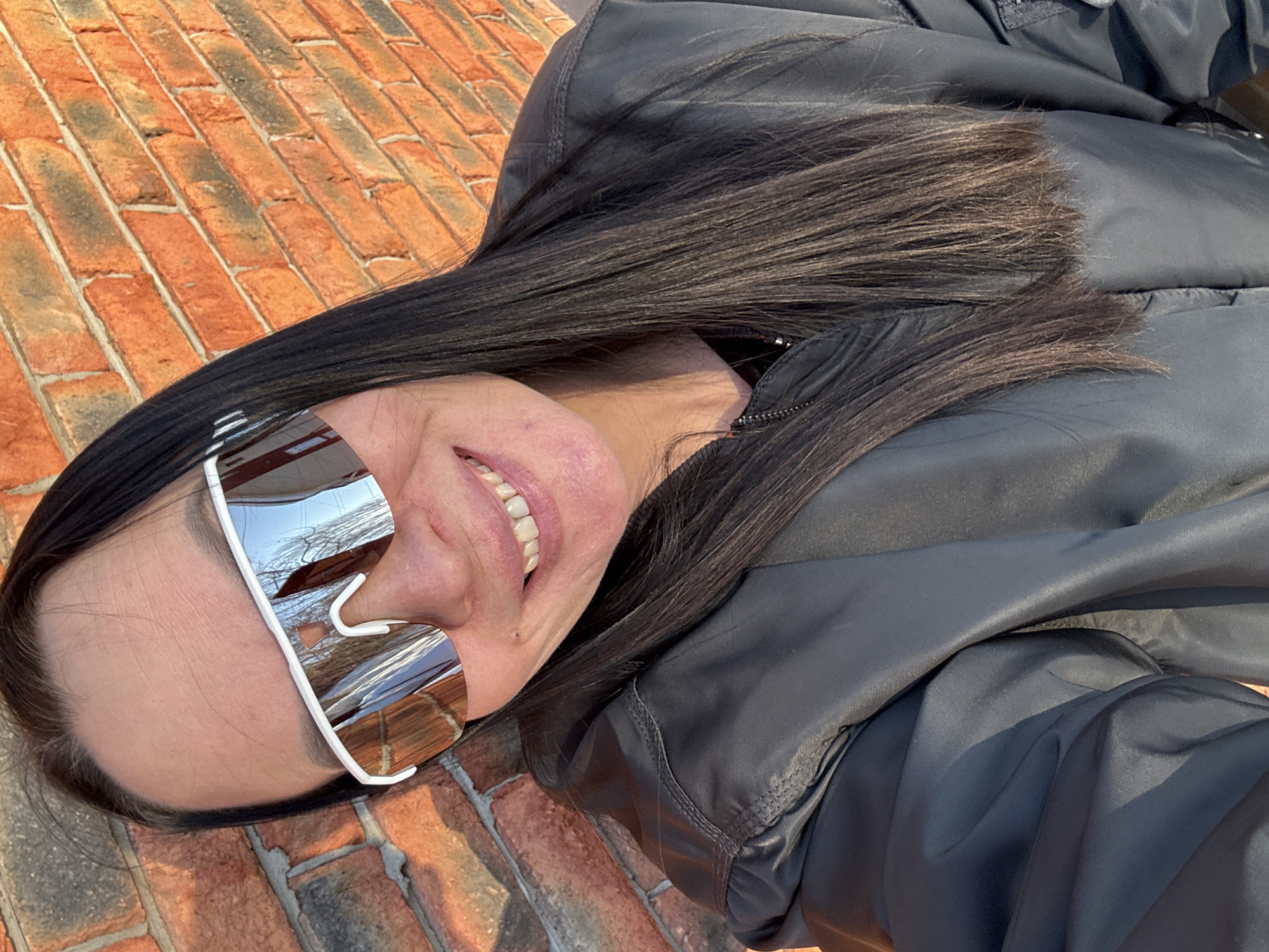 An adult female is depicted in a medium-close shot, positioned outdoors in front of a red-orange brick wall, in the city of Chaikovsky, Russia. She has long, straight, dark brown hair that falls over her shoulders. Her face is turned towards the camera, and she is smiling, revealing her upper teeth. She is wearing white-framed, mirrored shield sunglasses which reflect a clear blue sky and bare tree branches. Her attire includes a dark grey or black bomber-style jacket with a zippered front and a zippered utility pocket on the left arm, featuring visible stitching details. The brick wall behind her is illuminated by strong, direct sunlight, creating a warm hue on the bricks and some slight shadows.