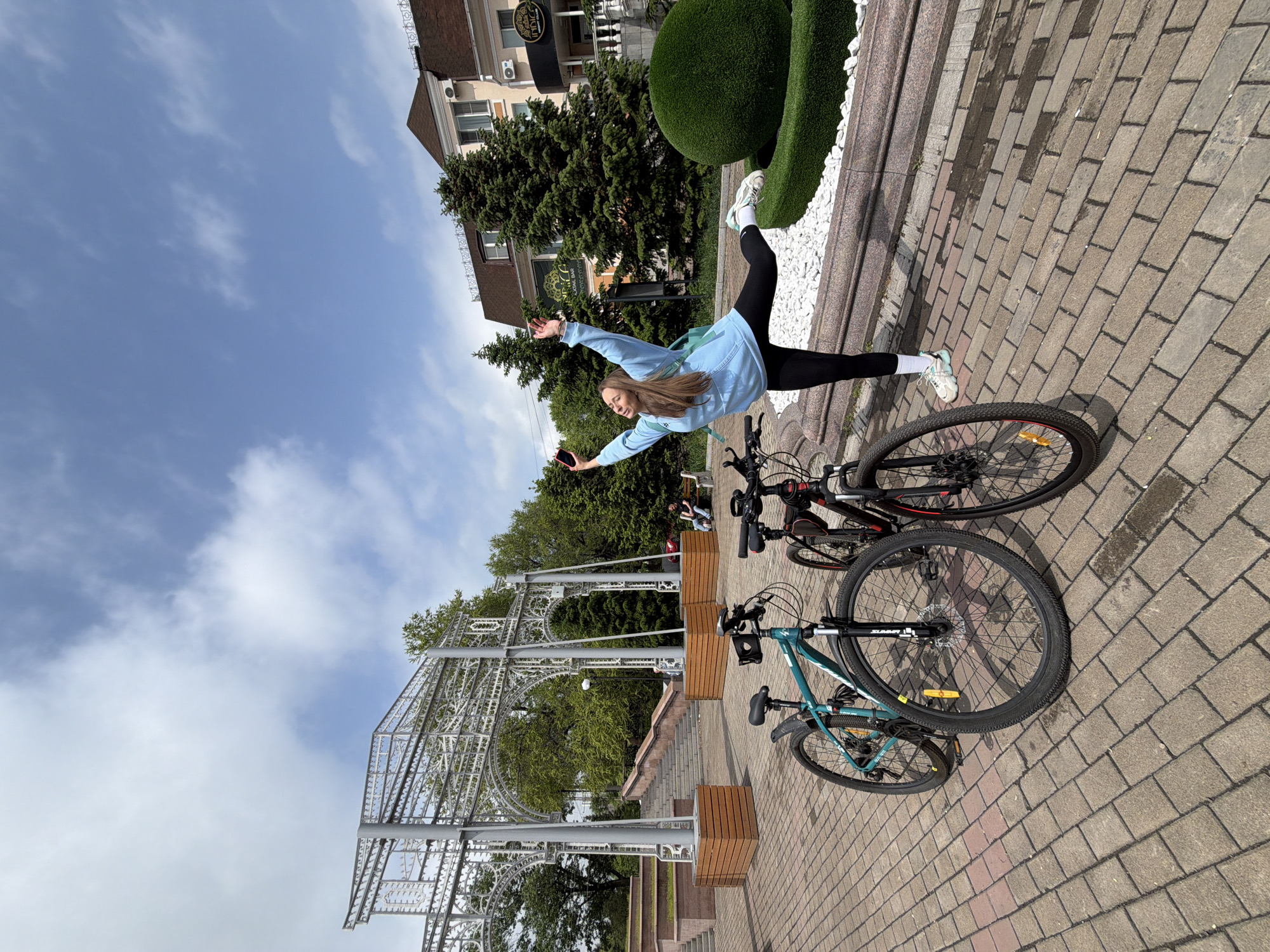 A young woman is positioned in a sideways horizontal split over two bicycles on a brick-paved area. She is wearing black leggings, white athletic shoes, and a light blue sweatshirt. Her arms are outstretched. She appears to be holding a phone in her right hand. The bicycles are parked side by side. A decorative white metal archway is visible in the background. Behind the woman and the bicycles, there is a raised platform with sculpted green shrubbery and a building in the background with a sign in Russian. The sky is blue with some clouds. Based on the sign and architectural style, the location is Birobidzhan, Russia.