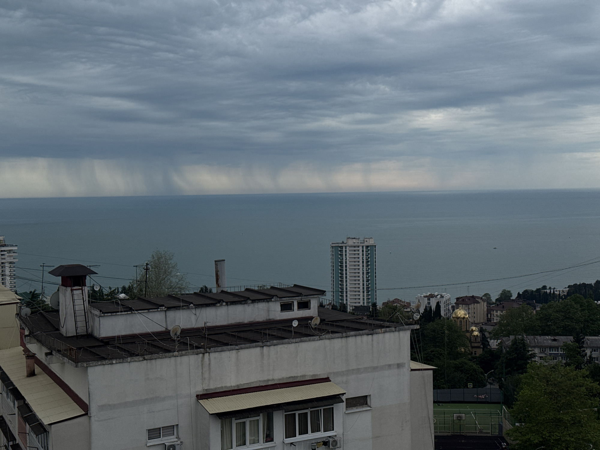 The image presents an overcast view of Sochi, Russia, overlooking the Black Sea. The scene is dominated by a sky filled with dense, grey clouds, with visible rain shafts descending into the ocean. A white, multi-story building with a flat, cluttered roof occupies the foreground. Various antennas, vents, and a ladder are seen on the rooftop. In the midground, a modern high-rise building stands near the coast, and a golden-domed Russian Orthodox church is visible among the trees. A sports field with a basketball hoop is also visible. The overall environment suggests a humid, possibly stormy, weather condition.
