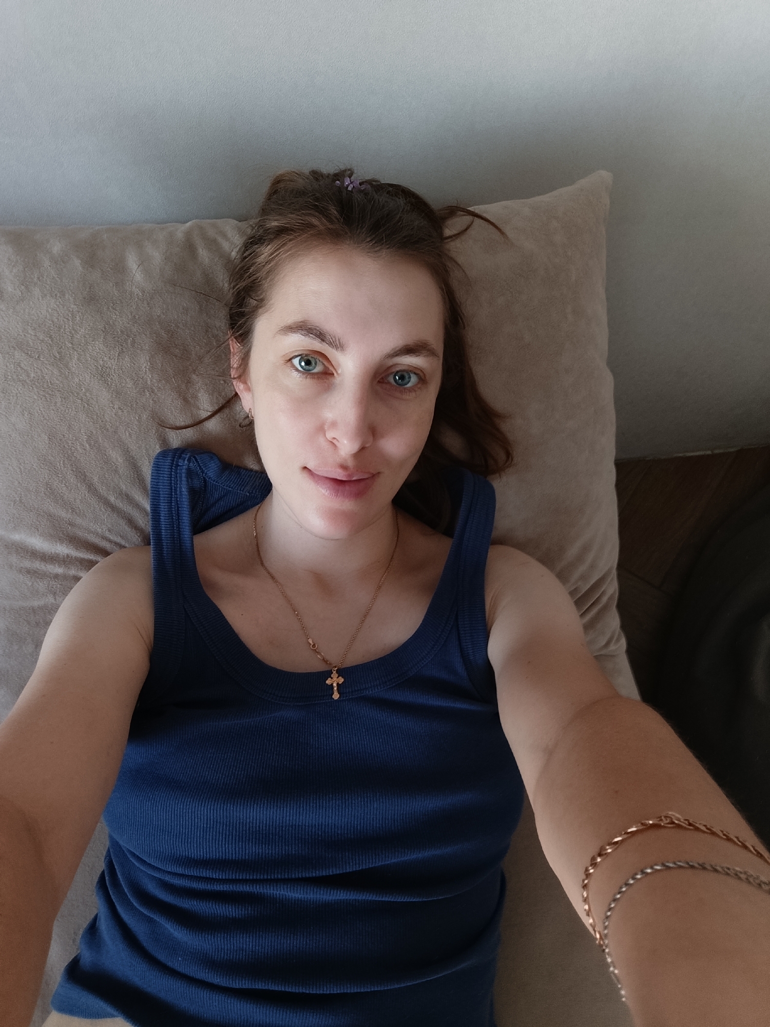 A young woman lies on a beige pillow, wearing a blue tank top and a gold necklace with a cross pendant. She is looking directly at the camera. She has light skin, brown hair, and blue eyes. Two bracelets are visible on her right arm. The background consists of a light gray wall and part of a brown sofa. The photo appears to be taken from above. The city is not discernable from this image.