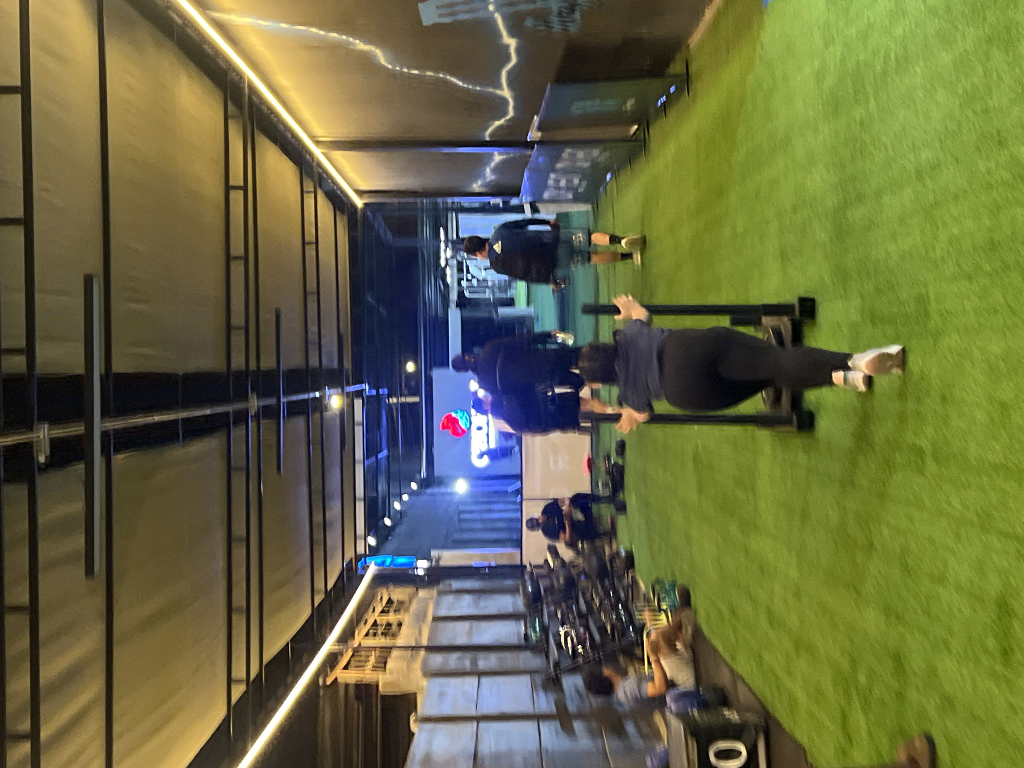 The image shows a gym setting in Arica, Chile. There are multiple people, with one individual prominently positioned pushing a sled on a green, artificial turf surface. Other people are present, either engaging in other exercises or resting. Gym equipment, including weights and benches, are visible. The gym has a semi-outdoor design with a metal frame structure and lighting fixtures. A sign with the word "Crossfit" is illuminated in the background.