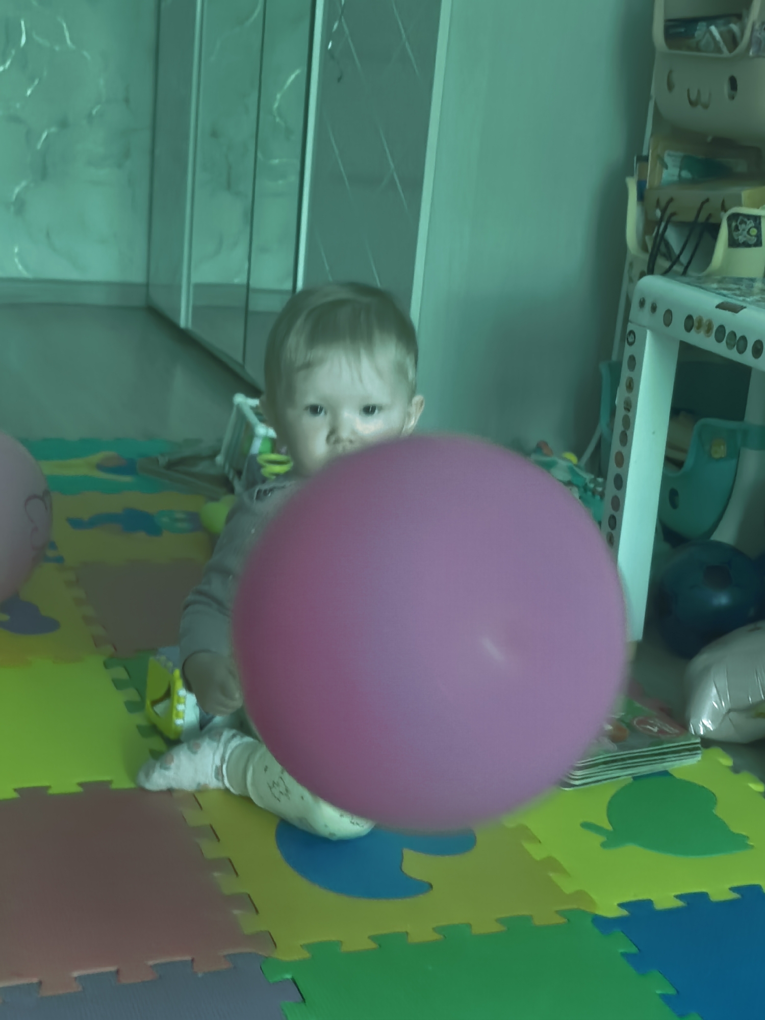The image captures a baby or toddler sitting on a colorful foam play mat indoors, looking towards the camera. The child has light-colored hair and is wearing a light-colored top and patterned socks. A large, bright pink balloon dominates the foreground, partially obscuring the baby's body and lower face. Another lighter pink balloon is visible on the left.
The floor is covered with interlocking foam play mats in various colors, including yellow, green, blue, brown, and red-orange. In the background, to the right, there is a white and beige multi-tiered storage unit or shelf, and part of a white plastic table or chair structure. A dark blue spherical object (possibly a toy ball) and a metallic silver-colored balloon or soft toy are also visible on the right. The room has a somewhat cool, greenish tint, especially in the background wall areas, which appear to have reflective or patterned surfaces.
The activity taking place is playtime, with the baby sitting amidst toys and play mats, seemingly engaged with the large pink balloon. The baby appears calm. No specific time of day or weather conditions can be determined from the indoor setting. There is no visible text in the image.