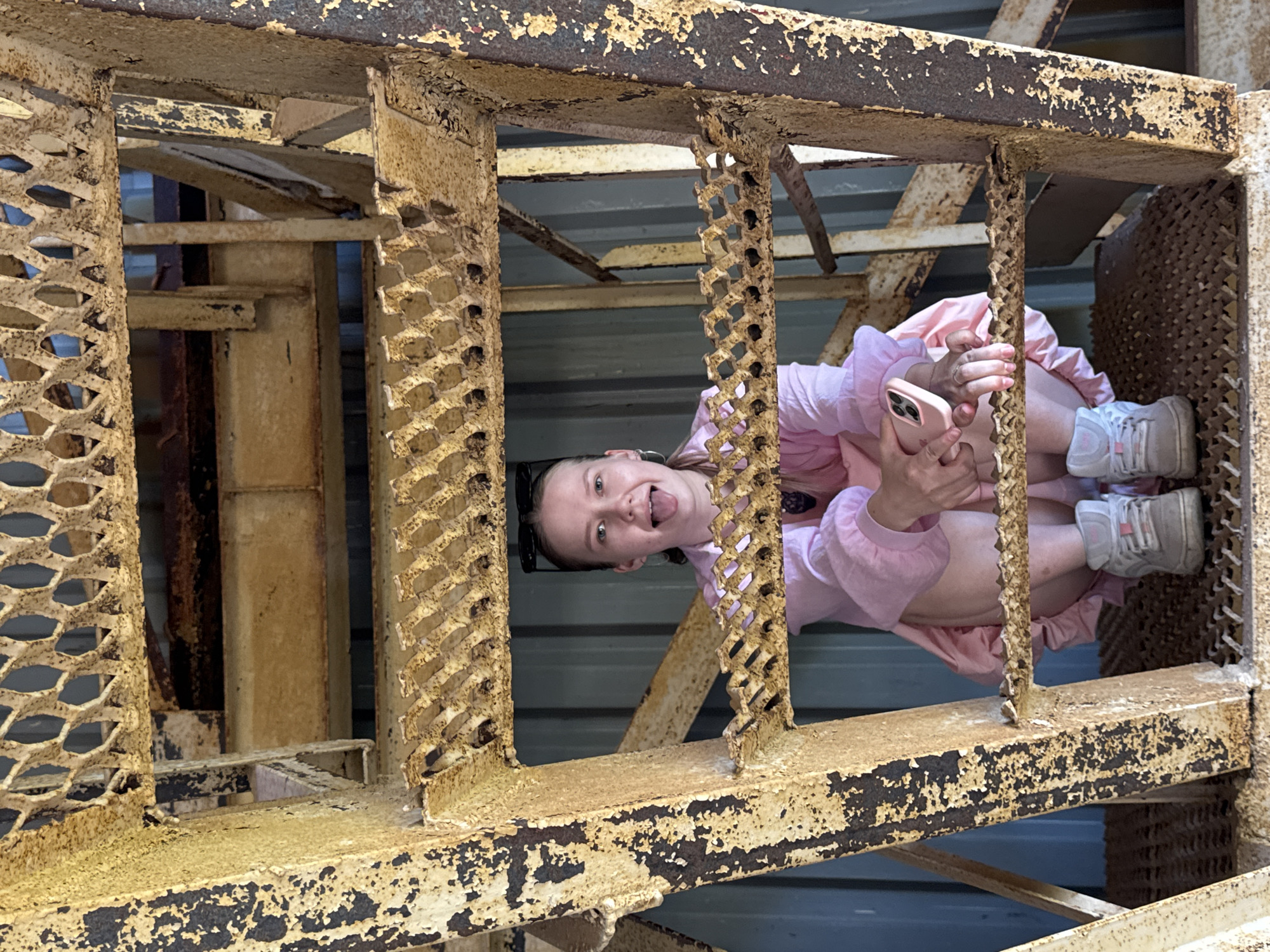 A young person with fair skin is seen upside down, framed by rusted metal structures. She has dark hair, and is wearing sunglasses on her head. She is sticking out her tongue. She is wearing a pink jacket and a pink dress with white sneakers. She is holding a pink phone. The location is Rusalevka, Russia, inferred from image source. The environment includes metallic, industrial-style beams and grating.