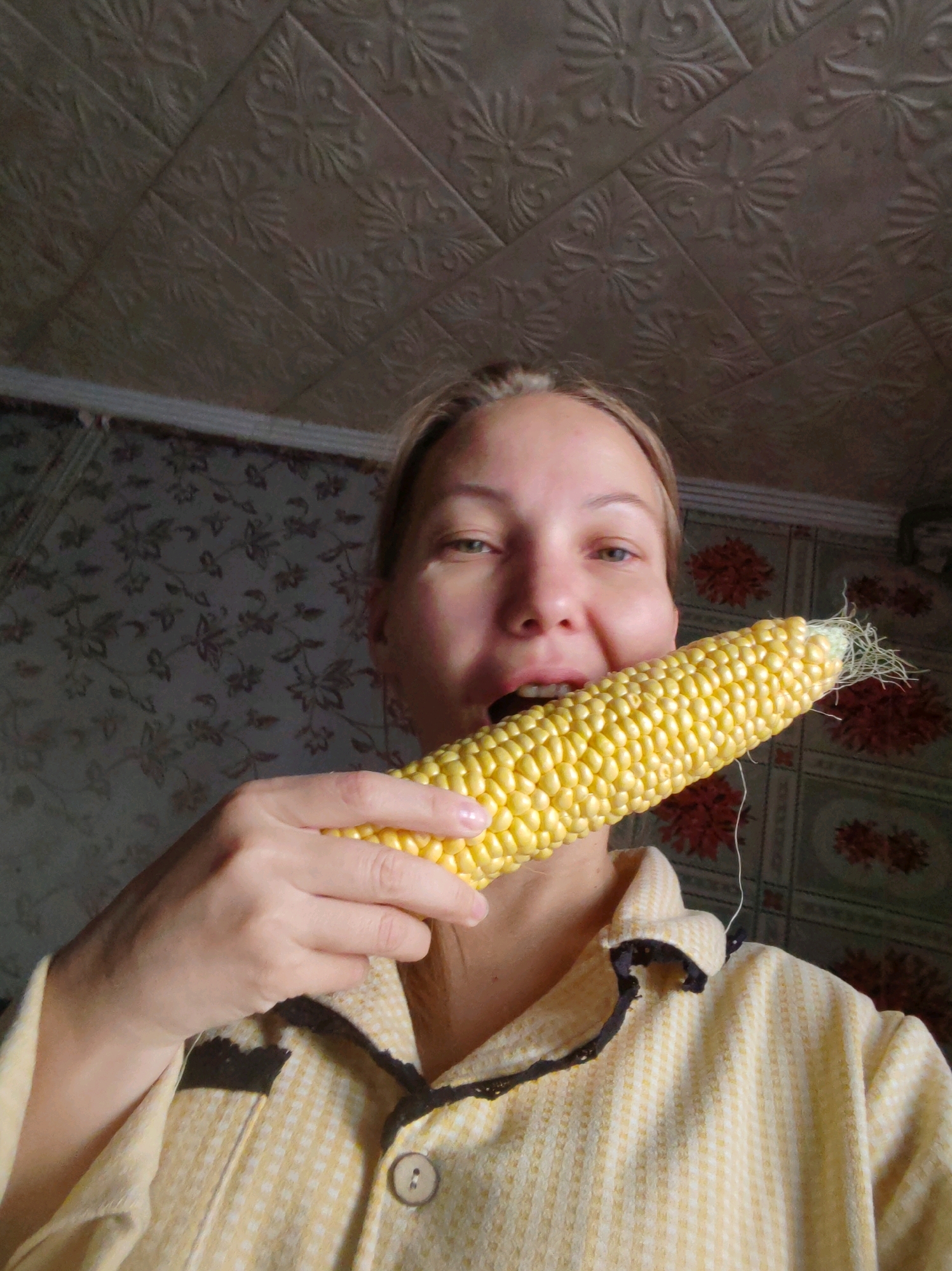 A woman with blonde hair pulled back is shown from the chest up, holding a corn cob to her mouth. She is wearing a yellow plaid pajama top with a black trim. The background appears to be a room with patterned wallpaper and a textured ceiling. The lighting is dim and natural. The woman's face is partially obscured by the corn cob.
