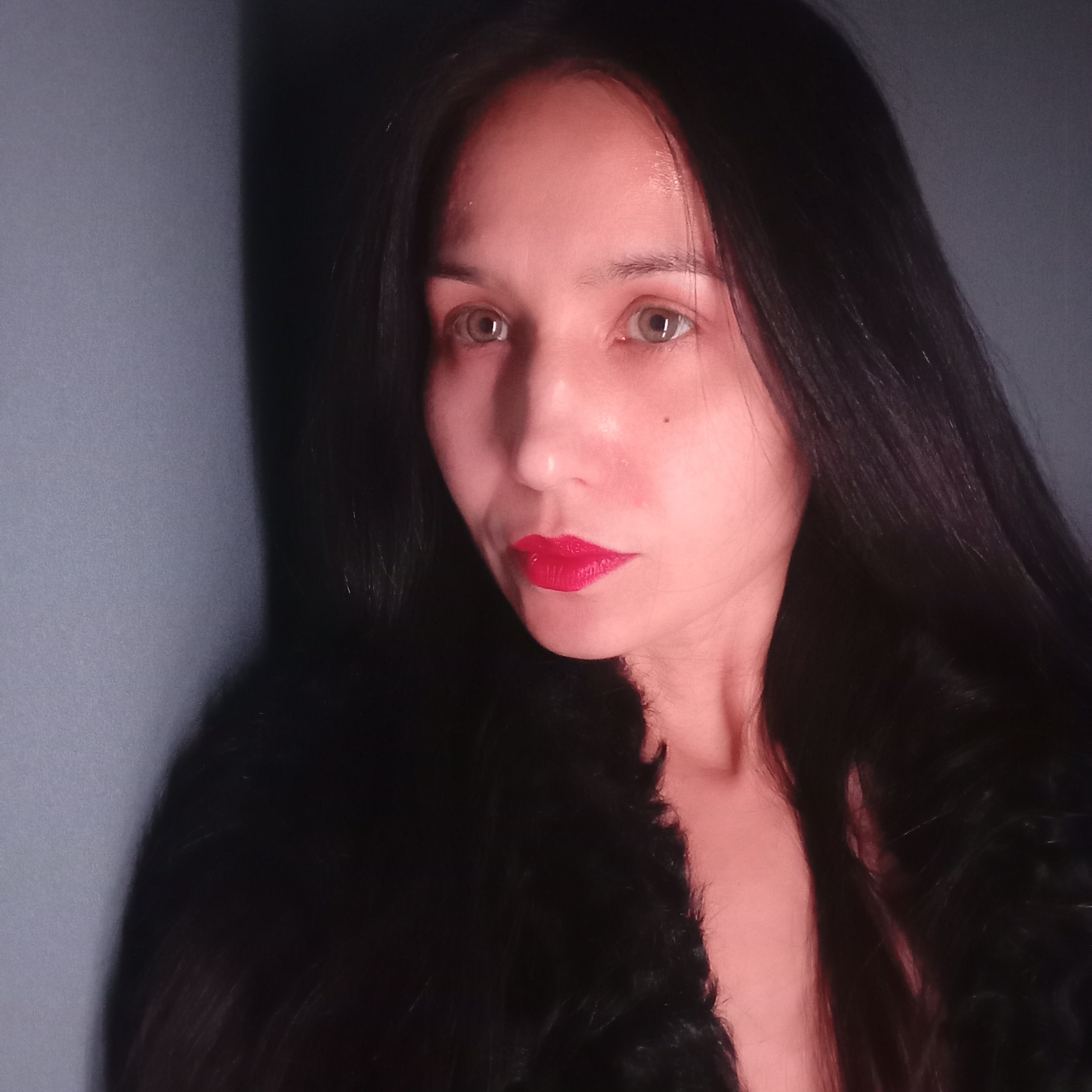 A woman is in a dimly lit indoor setting with a blue-gray wall in the background. She has long, black hair, hazel eyes, and is wearing bright red lipstick. She appears to be wearing a dark, possibly fur, garment. There is no indication or implication that this image was taken in Pervomaysky, Russia or anywhere else.