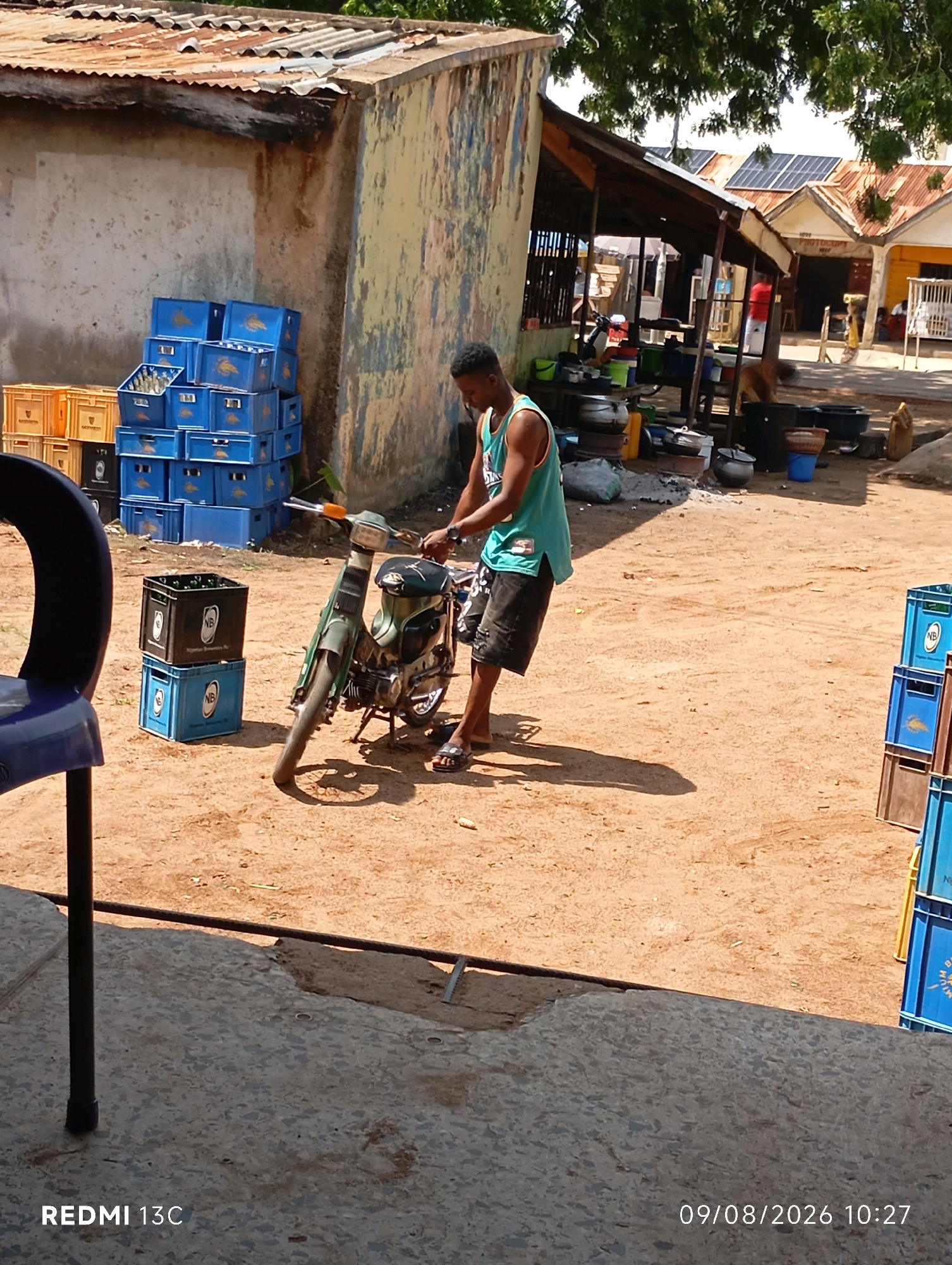 The image depicts an outdoor scene in Shendam, Nigeria, likely in a market or roadside area. A young man, wearing a teal tank top and black shorts, is standing next to a green motorcycle. He is holding onto the handlebars of the motorcycle, which is propped up on its stand. The motorcycle appears to be an older model.
In the background, there are several stacks of blue and orange plastic crates, some of which contain bottles. These crates are typical for transporting beverages. Adjacent to the crates is a partially dilapidated building with a corrugated metal roof and weathered walls, suggesting a utilitarian and possibly impoverished environment. Further in the background, other structures with corrugated roofs are visible, and some appear to have solar panels, indicating a mix of traditional and modern infrastructure. The ground is a sandy, dirt surface, common in many parts of Nigeria. The overall impression is that of a busy, functional, and somewhat rustic commercial area.