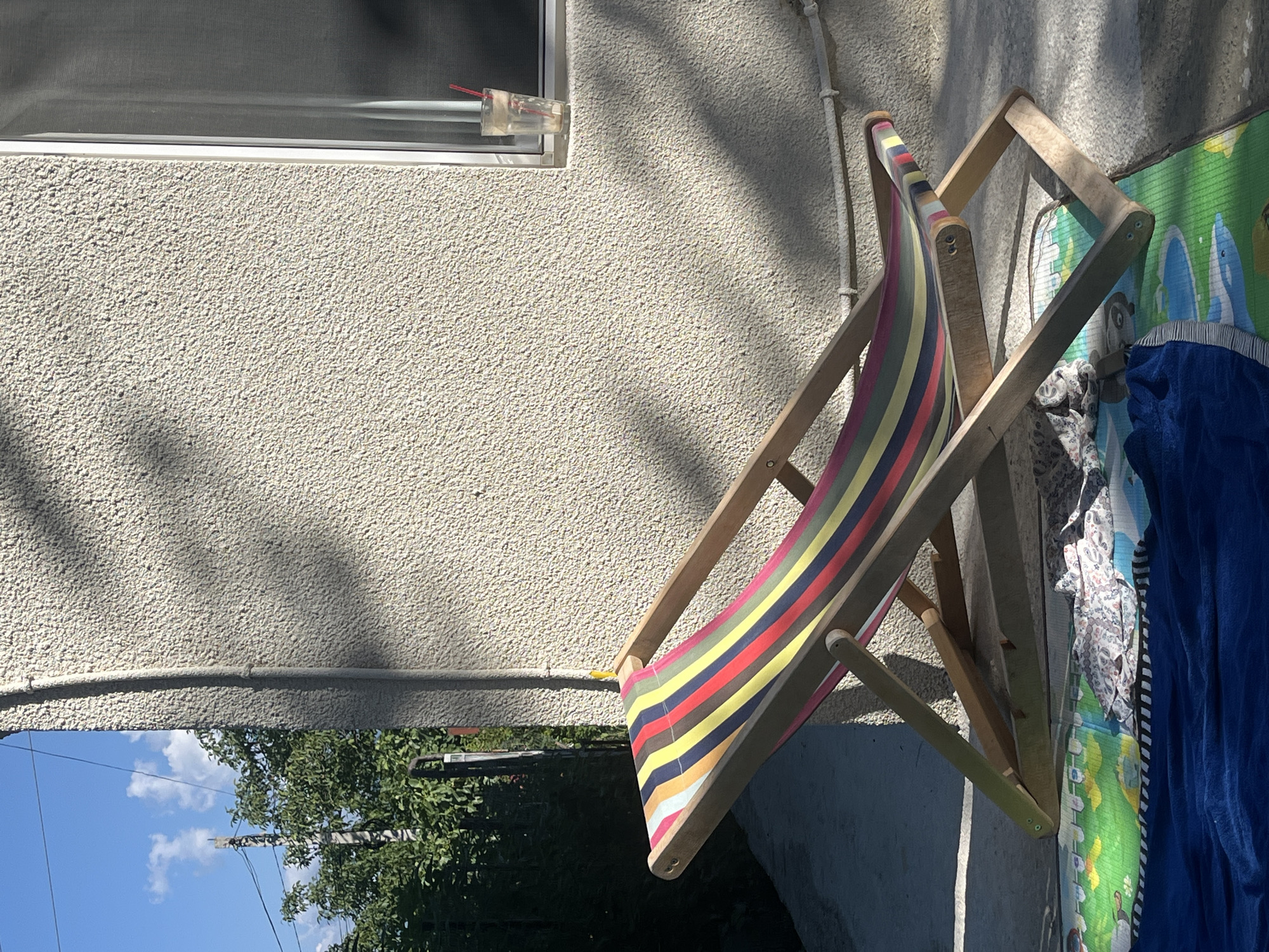 This image shows a striped deck chair leaning against a textured, pale wall. A window with a screen is visible on the wall, and a clear plastic cup with red straws sticks out of the window. Sunlight casts shadows on the wall, suggesting it is daytime. Beside the deck chair is a blue blanket and a colorful playmat with cartoon characters on it. Some floral fabric is draped over the deck chair's frame. In the background, the sky is blue with white clouds, and green trees are visible. The scene appears to be outdoors, possibly in a yard or on a patio. There is no visible text.