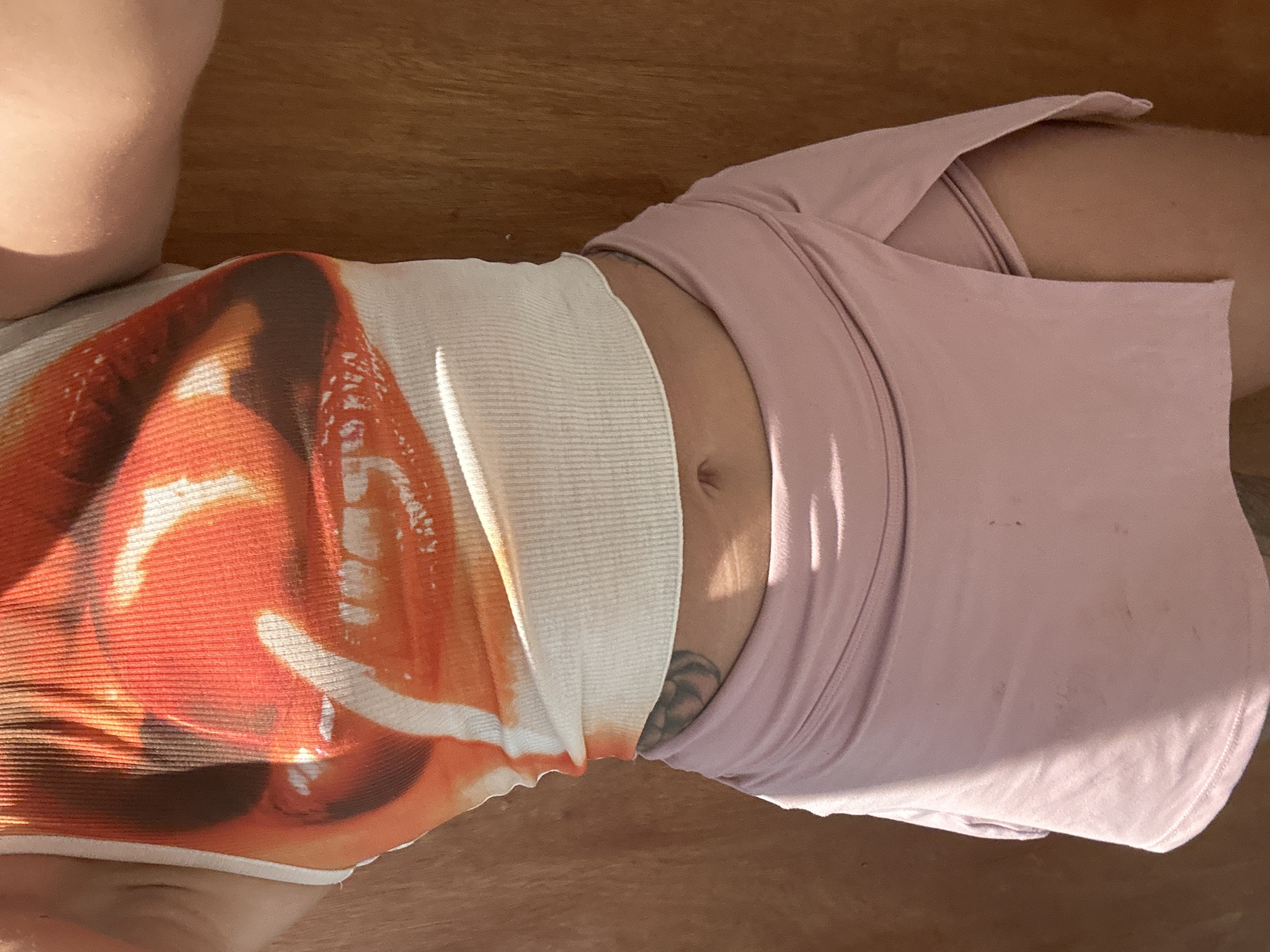 A person stands against a wooden background, their midriff exposed. They are wearing a cropped, ribbed tank top with a bold, abstract print of orange and red lips. Below the tank top, they wear a pink wrap skirt. A tattoo is visible on their lower torso, partially covered by the skirt. The lighting suggests an outdoor setting, casting shadows and highlighting the textures of the clothing and background. The location is unspecified.