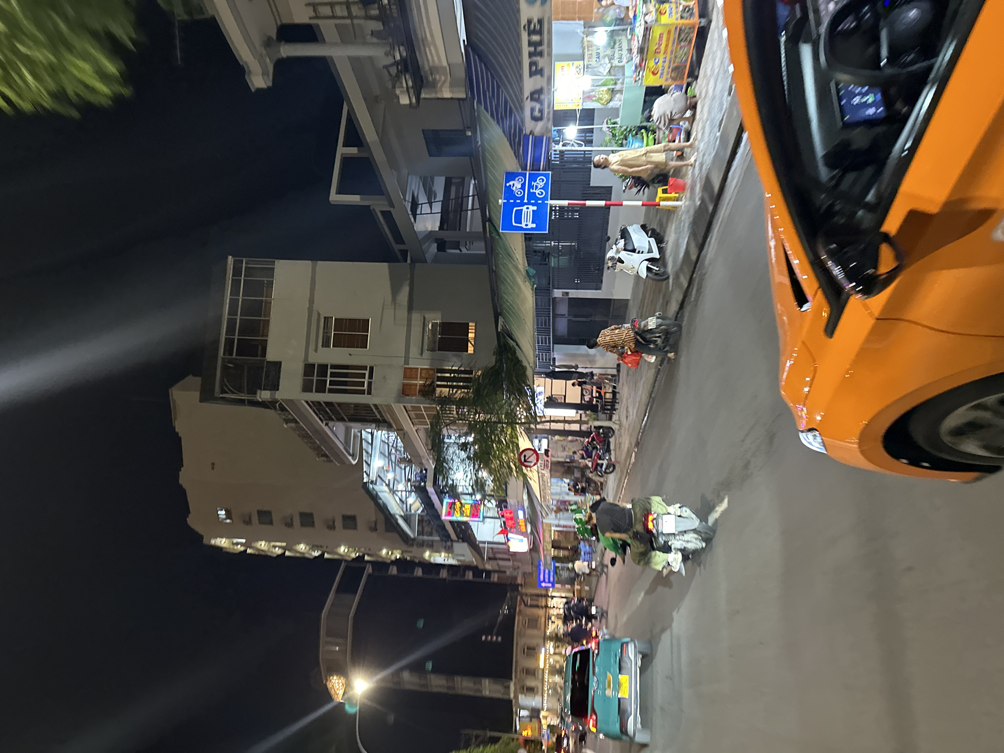 The image captures a street scene at night in Nha Trang, Vietnam, likely on a commercial street with multi-story buildings lining the road. The perspective is from a vehicle in motion, looking forward and slightly to the right.
In the foreground, the bright orange paint of the vehicle the photo is taken from is prominent, showing part of the hood, windshield, and driver's side mirror.
The street is moderately busy with traffic. A light blue sedan is visible in the middle distance on the left, with its rear end facing the camera. Several motorcycles are also present. One motorcycle in the center of the frame has a rider wearing a green helmet and a person in the back, both dressed in dark clothing. Another motorcycle with a rider in a striped shirt is further down the road on the right. A white scooter is parked on the sidewalk.
The buildings along the street are illuminated by artificial light. Many have storefronts at ground level, with awnings and signs. One sign clearly reads "CÀ PHÊ S". Another sign displays Vietnamese text, possibly advertising food or drinks. A blue traffic sign with symbols for a car and two types of bicycles is posted on a pole.
On the sidewalk to the right, a woman in a light-colored dress stands near a food stall. Another person is crouched down, possibly attending to the stall. Various items are displayed at the stall, appearing to be food items.
The overall environment suggests a lively urban setting at night, with the sounds and sights of a bustling street. The lighting conditions are typical of a nighttime city environment, with streetlights and building illumination casting shadows and highlighting the scene. The presence of multiple vehicles, pedestrians, and commercial establishments points to a well-traveled and active area.