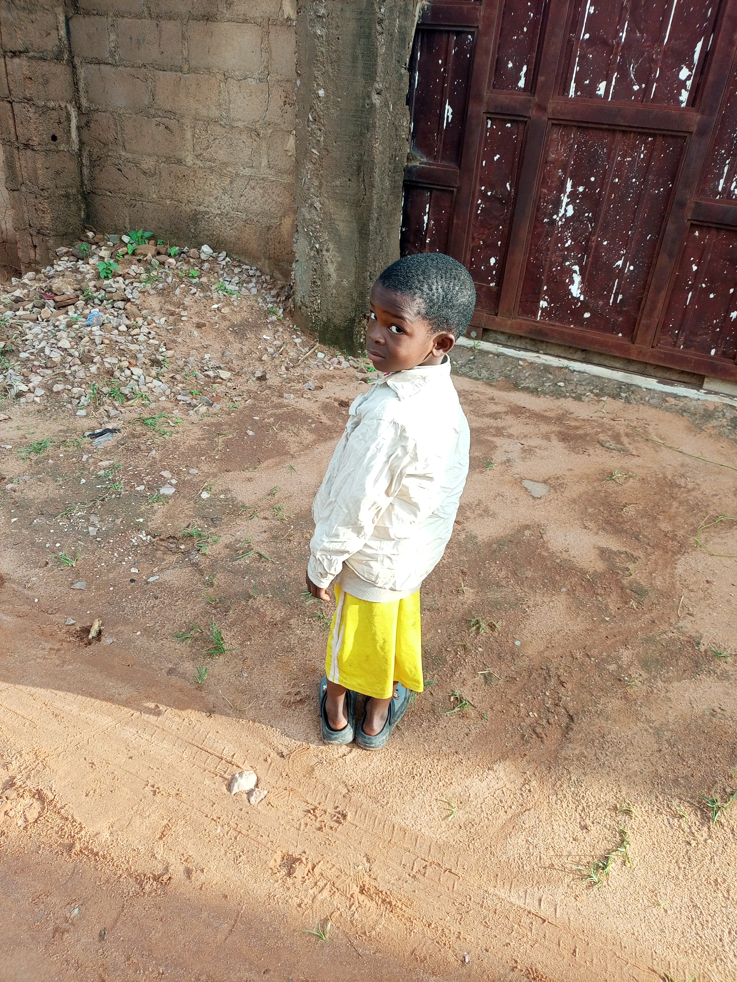 In Gombe, Nigeria, a young boy stands outside on a dirt ground. He is wearing a light-colored jacket and yellow pants. Behind him, a rough-hewn brick wall runs parallel to a large, dark brown metal gate with white paint splatters. The ground is covered in reddish-brown dirt, small rocks, and sparse patches of green grass. The lighting suggests it is daytime, with shadows cast on the ground.