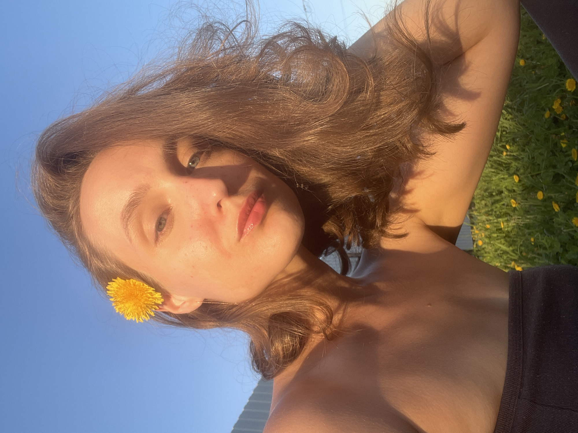 A young woman poses outdoors in Подшивалово, Russia. She has brown, wavy hair and is wearing a small yellow flower tucked behind her ear. Her skin is illuminated by sunlight, creating soft shadows. The background features a clear blue sky and a patch of green grass dotted with yellow flowers. Part of her black top is visible in the lower right corner of the frame.