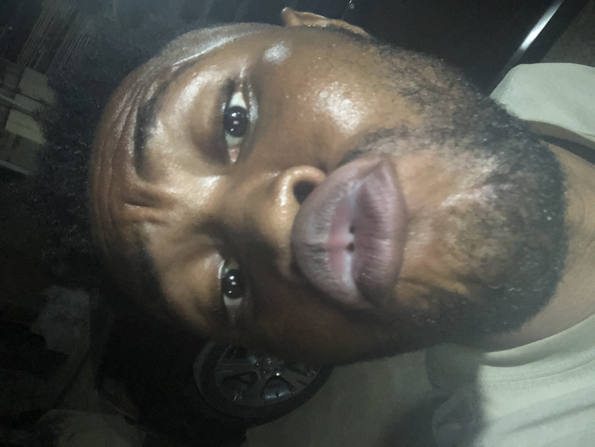 A close-up, slightly angled selfie of a Black male with dark eyes, short dark hair, and a trimmed beard. His skin appears slightly shiny, possibly due to moisture. He is pursing his lips. Part of a light-colored shirt is visible at the lower right edge. The background is dark and indistinct, with a partially visible object resembling a car wheel towards the lower center. The location is Unknown, Unknown.