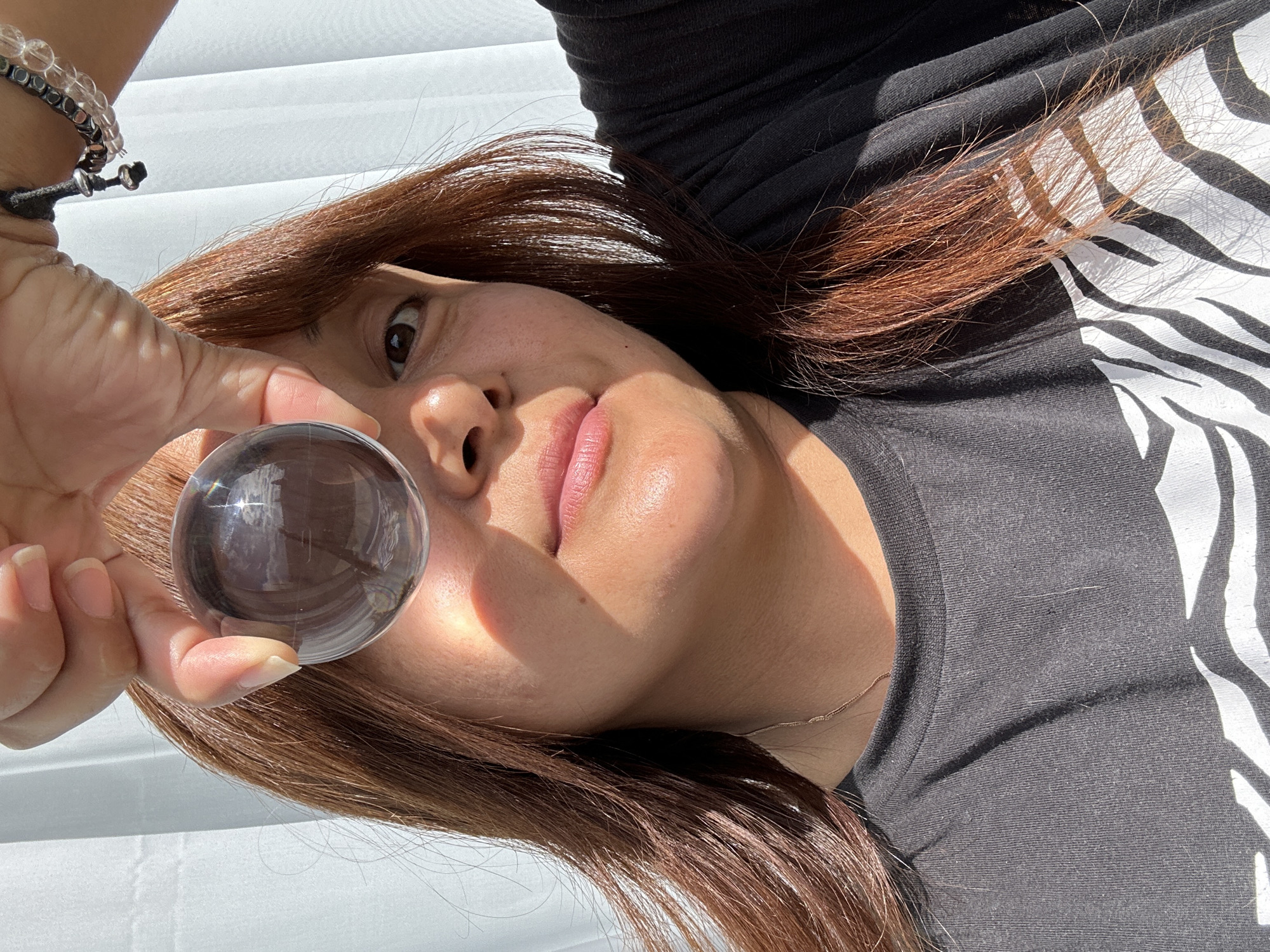 An individual is depicted in a close-up, eye-level portrait, holding a clear, spherical crystal ball in their right hand, positioned in front of their right eye. The individual has a tan complexion with visible skin texture, and long, reddish-brown hair is draped over their shoulders. Their visible left eye is brown. A thin, gold-colored necklace is partially visible around their neck, and two bracelets adorn their right wrist: one dark-colored with a silver-toned bead, and another composed of clear, spherical beads. They are wearing a black, short-sleeved t-shirt featuring a white, zebra-stripe-like graphic on the lower right portion. Strong, directional natural light originates from the viewer's right, casting distinct highlights on the left side of the individual's face, nose, and lips, and creating shadows on the right side of the face and neck. The crystal ball refracts and inverts the background, revealing distorted elements of light-colored, vertically pleated curtains, which serve as the backdrop. The scene is located indoors in Unknown, Unknown.
