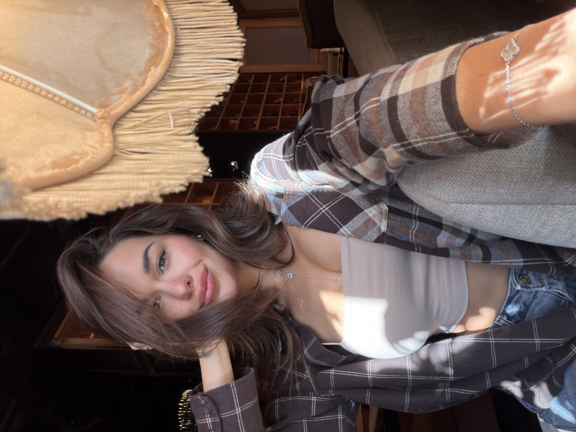 A young woman is posing for a photo. She is wearing a brown and tan plaid shirt, a white tank top, and blue jeans. She is also wearing a silver necklace and a bracelet. She is resting her head on her hand. A lamp with a beige lampshade and fringe is partially visible in the upper left corner. A shelf filled with small objects is visible in the background.
It is not possible to determine if the photo was taken in Krasnoyarsk, Russia, from the image alone.