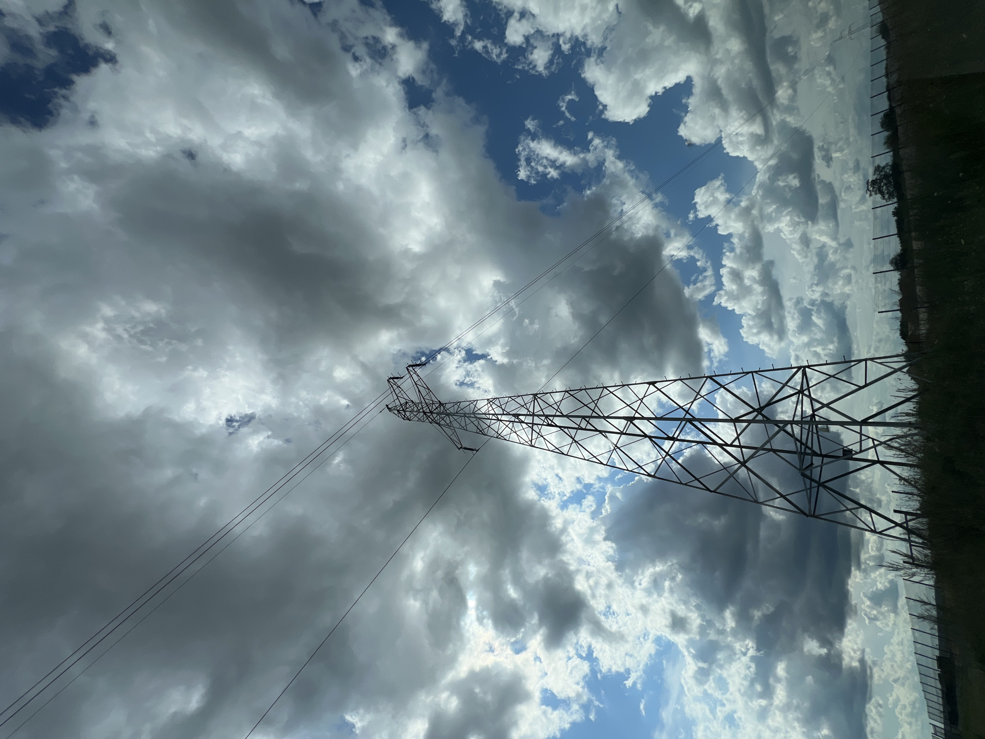A tall, metal electricity pylon stands in silhouette against a dramatic sky filled with both dark, heavy clouds and patches of bright blue. Power lines extend diagonally from the top of the pylon and disappear towards the upper left of the frame. The pylon's lattice structure is clearly visible, with a dark green, grassy field and a low fence in the foreground. The scene appears to be an outdoor, rural or semi-rural location, possibly on the outskirts of Fasty, Poland, given the power infrastructure and open landscape.