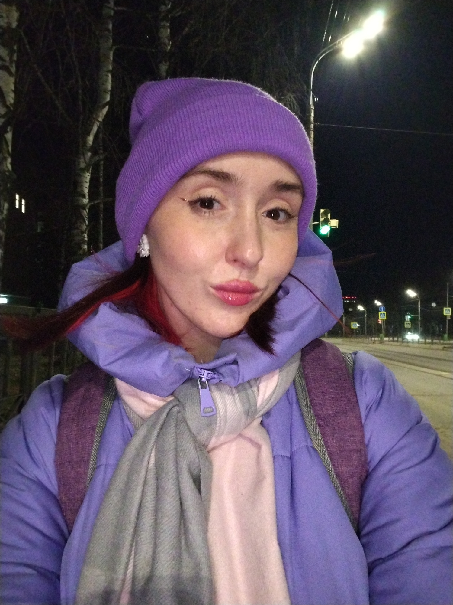 A fair-skinned female wearing a purple beanie, purple hooded winter coat, and a scarf is standing on the sidewalk in Izhevsk, Russia, at night. She is wearing earrings and has red streaks in her hair. Her lips are pursed.
Behind her is a street with multiple street lights. Road signs are visible. Trees are to the left.