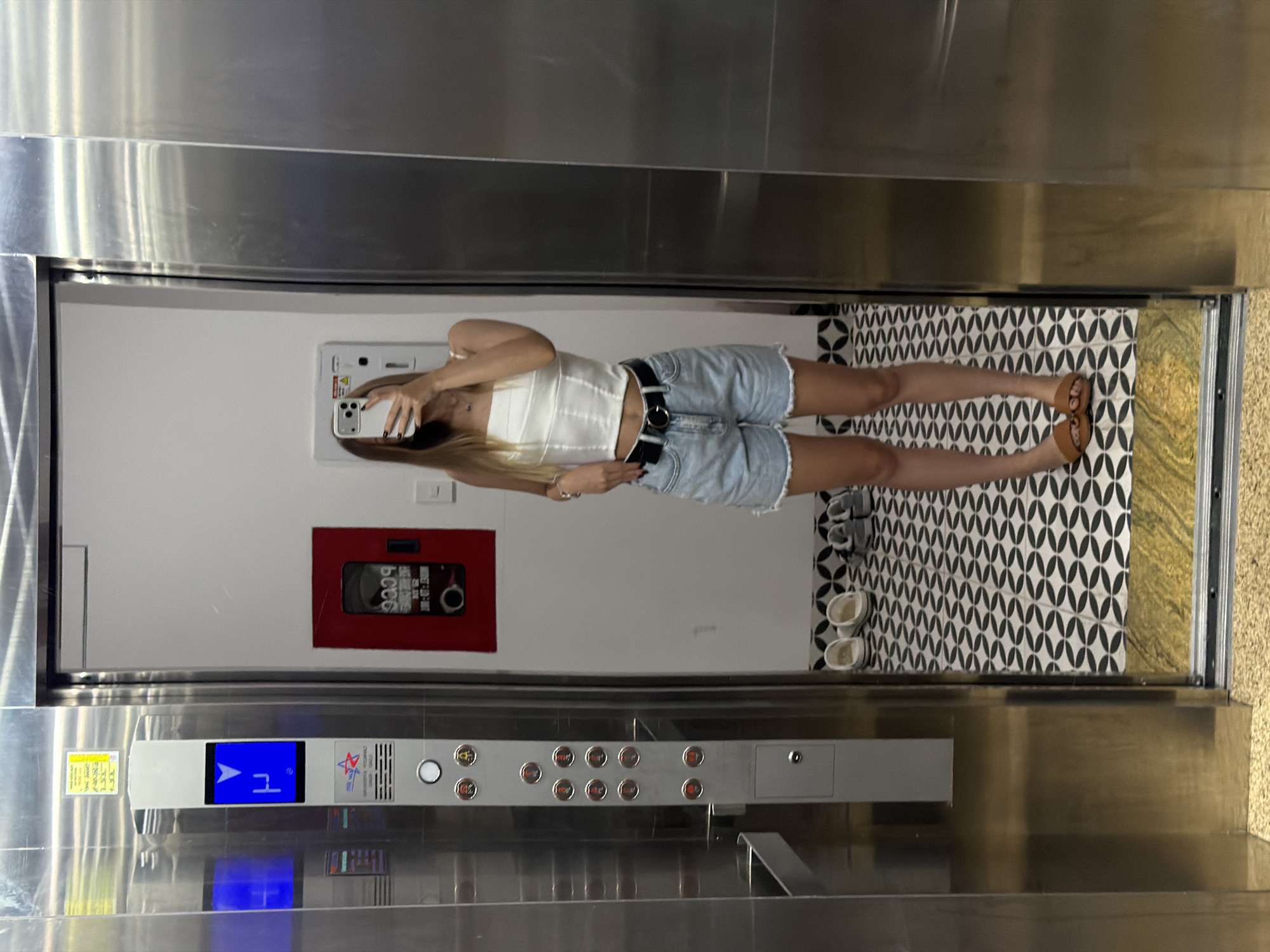 A young woman stands in an elevator with its doors open, holding a smartphone to take a selfie. She is wearing a white corset top, denim shorts, a black belt with a gold buckle, and brown sandals. The elevator interior features stainless steel walls and an elevator control panel with illuminated buttons. The floor inside the elevator is tiled with a black and white geometric pattern. Outside the elevator, in the hallway, there are a few pairs of shoes on the floor, and a red fire extinguisher box mounted on the wall. The image appears to have been taken in an urban setting, likely within a building in the city of Đà Nẵng, Vietnam, given the context.