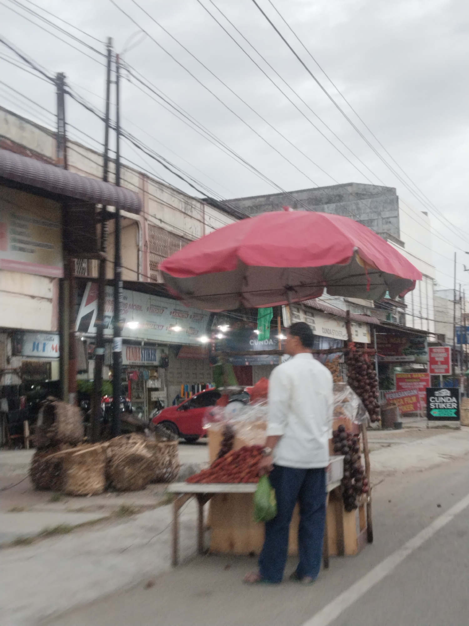 The image depicts a street scene in Lhokseumawe, Indonesia, focusing on a street vendor under a large red umbrella. A man, likely the vendor, wearing a white long-sleeved shirt and blue pants, stands near a display of goods. In front of him is a table laden with what appear to be fruits and possibly other packaged items. To his right, a rack holds several bunches of what could be rambutans or a similar fruit, along with some other produce. Woven baskets are stacked on the ground to the left of the vendor. Behind the vendor and the stall, a red car is visible, and further back, storefronts line the street. Overhead, a network of power lines crisscrosses the cloudy sky. The overall impression is of a bustling commercial area on an overcast day.