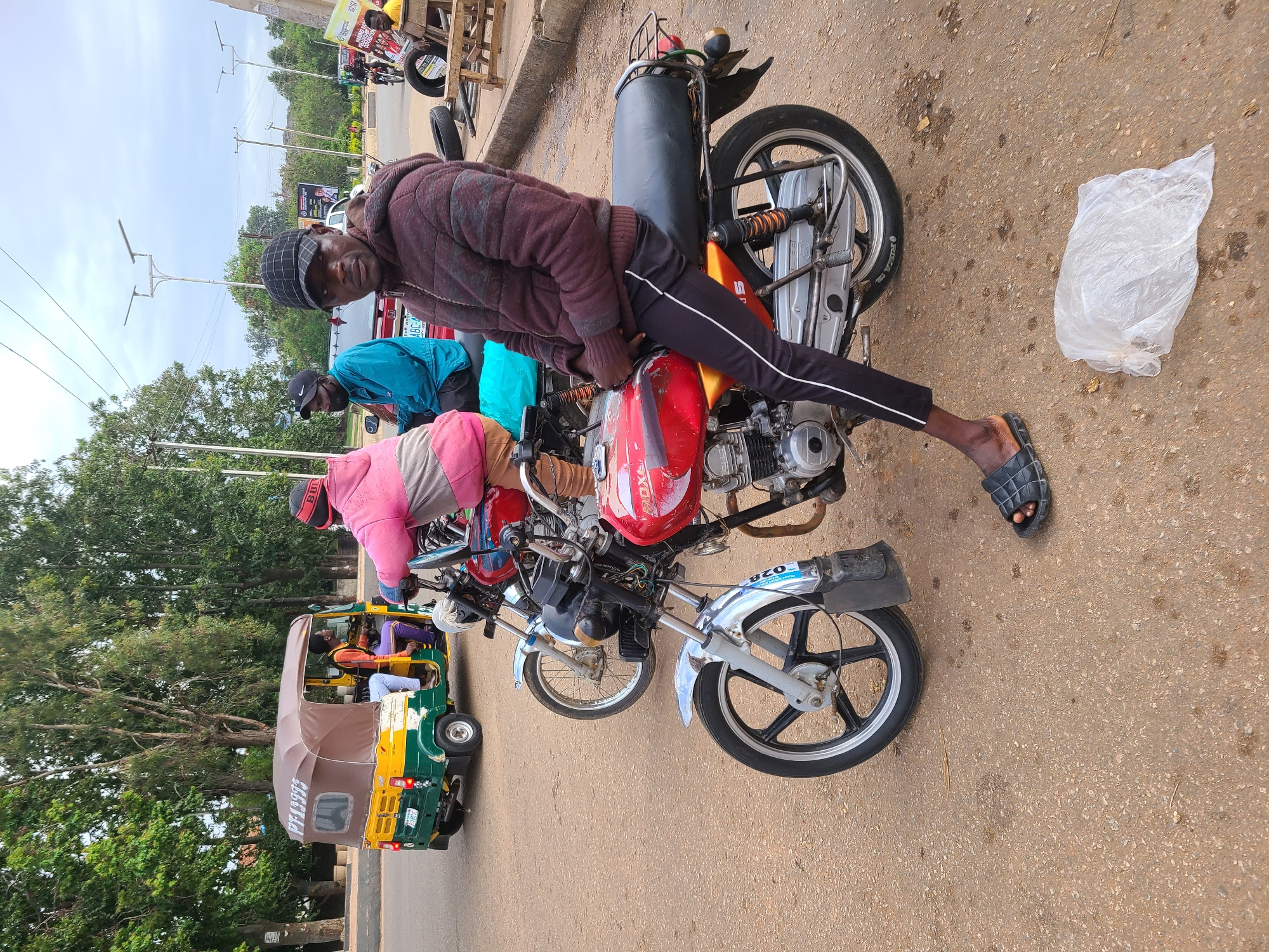 The image shows a man sitting on a parked motorcycle on a paved road in Jos, Nigeria. The motorcycle is red and silver with a black seat. The man is wearing a dark-colored jacket, black pants, and a cap. He is looking at the camera. In the background, there are other people, motorcycles, and an auto rickshaw (tricycle). A clear plastic bag sits on the road near the motorcycle. The sky is overcast and there are trees and buildings in the background.